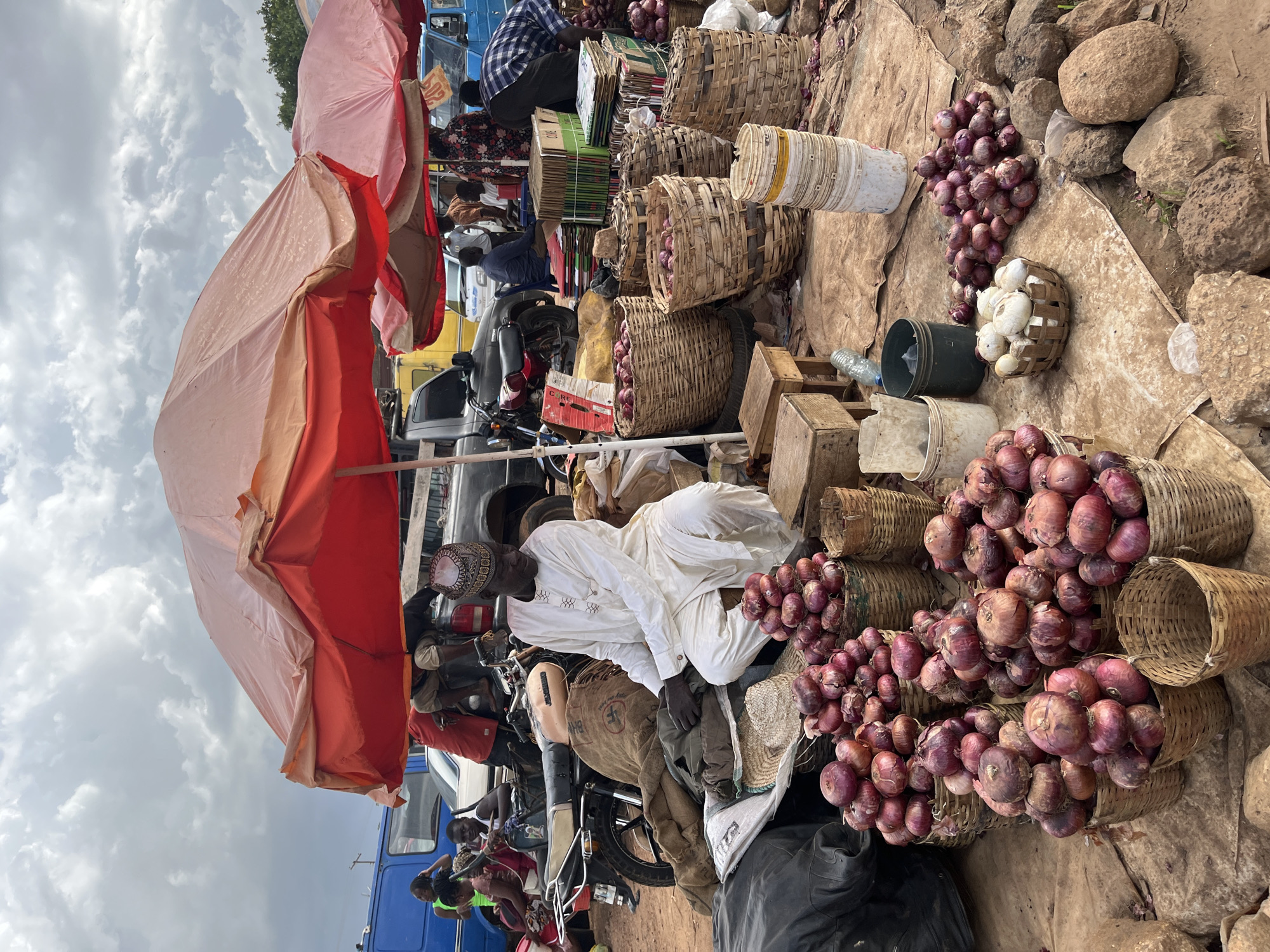 In Rafin Par, Nigeria, a man in traditional white attire with a patterned cap sits outdoors at a market stall, tending to a large display of red onions. The onions are piled in baskets and on the ground, some in woven baskets of various sizes. Behind him, several other people are visible, some interacting with vehicles and other market stalls. A motorcycle is parked to his left, with a burlap sack resting on it. Various umbrellas, one a prominent red and orange, provide shade. In the background, multiple vehicles, including a blue bus, a yellow truck, and a black pickup truck, suggest a busy marketplace environment. The ground is covered with a brown tarp and scattered debris. The sky is cloudy, indicating a potentially overcast day.