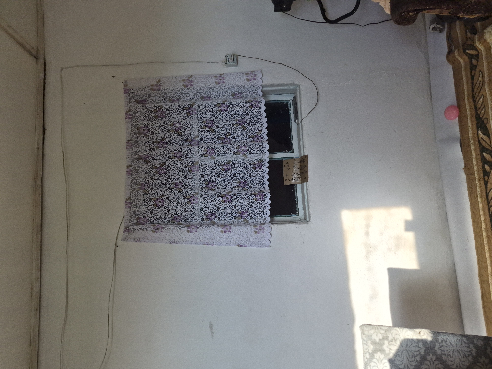 The image depicts an interior scene, possibly a room with a window. The walls are painted white, and there are some electrical wires running along the ceiling and down the wall. A window is covered with a white lace curtain with a floral pattern in purple and green. The window itself has a simple frame. Attached to the outside of the windowpane is a small piece of paper, possibly a note or advertisement. Sunlight is casting a bright rectangle of light onto the wall to the left of the window, and also onto a portion of a patterned sofa visible in the lower left corner. A section of a patterned rug is visible on the floor. In the lower right corner, a portion of a dark, upholstered furniture item is visible, and on the floor near it is a small pink ball. The overall impression is of a simple, possibly rural interior. No specific location in the city of Қанабек, Kazakhstan can be determined from the image alone.