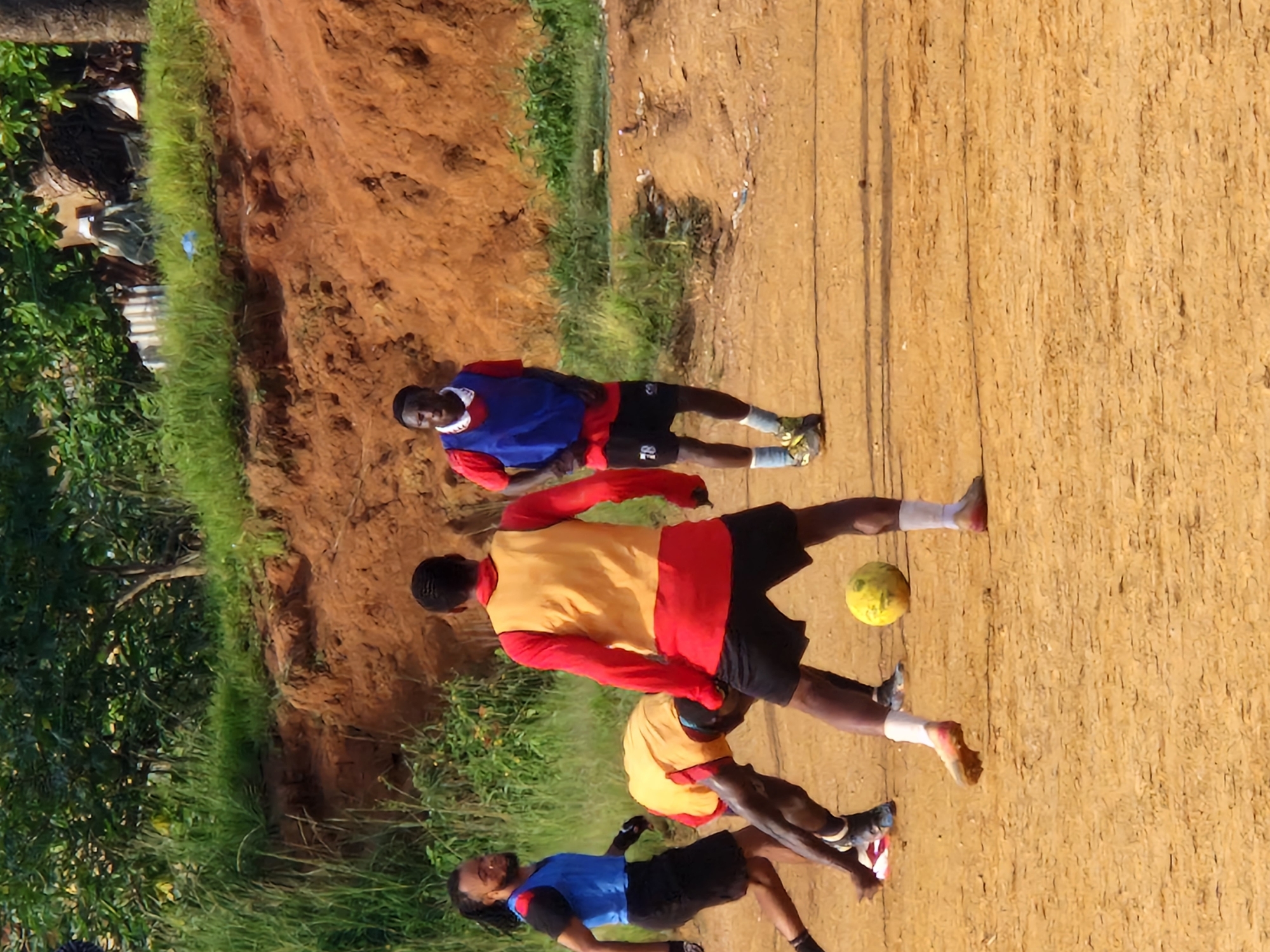 In Kabong, Nigeria, four young men are engaged in a football match on a dirt field. Two players, wearing blue vests over red shirts and black shorts, are actively participating in the game. One player, with a blue vest over a red shirt, is positioned in the foreground, facing left. Another player, wearing a blue vest over a red shirt and black shorts, stands in the middle ground. Two other players are also visible. One, in a yellow vest and red shirt, is bending over, appearing to interact with the ground. The fourth player, wearing a red long-sleeved shirt, is also in the middle ground, facing away from the camera. A yellow football rests on the ground in the foreground. The environment consists of a reddish-brown dirt field, with a steep, eroded embankment of the same color behind the players. Lush green vegetation covers the top of the embankment and extends into the background. Scattered debris is visible on the field near the embankment. The lighting suggests it is daytime, with shadows cast on the ground.