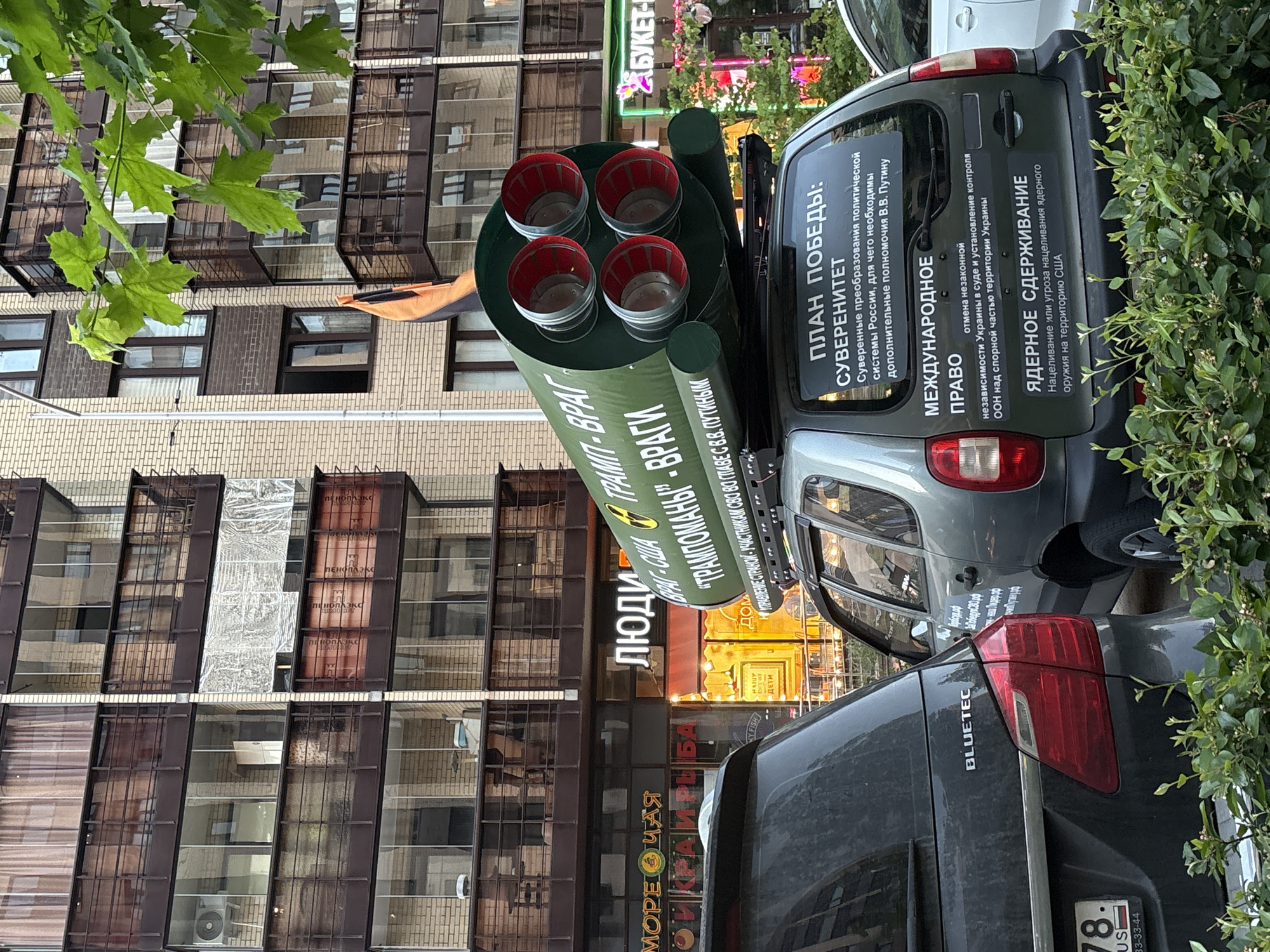 A dark grey Mercedes-Benz SUV, partially visible in the foreground, is parked next to a light grey Chevrolet Niva. Both vehicles are parked on a street in front of a multi-story brick apartment building. The Chevrolet Niva has a large, green cylindrical object mounted on its roof, resembling a rocket or missile launcher with four exhaust nozzles. The cylinder has text written on it in white Cyrillic letters. The rear window of the Chevrolet Niva also has several stickers with text in Cyrillic. The building behind the cars has multiple balconies with glass enclosures. The street appears to be paved. The scene is set in the city of Sinyavino, Russia.