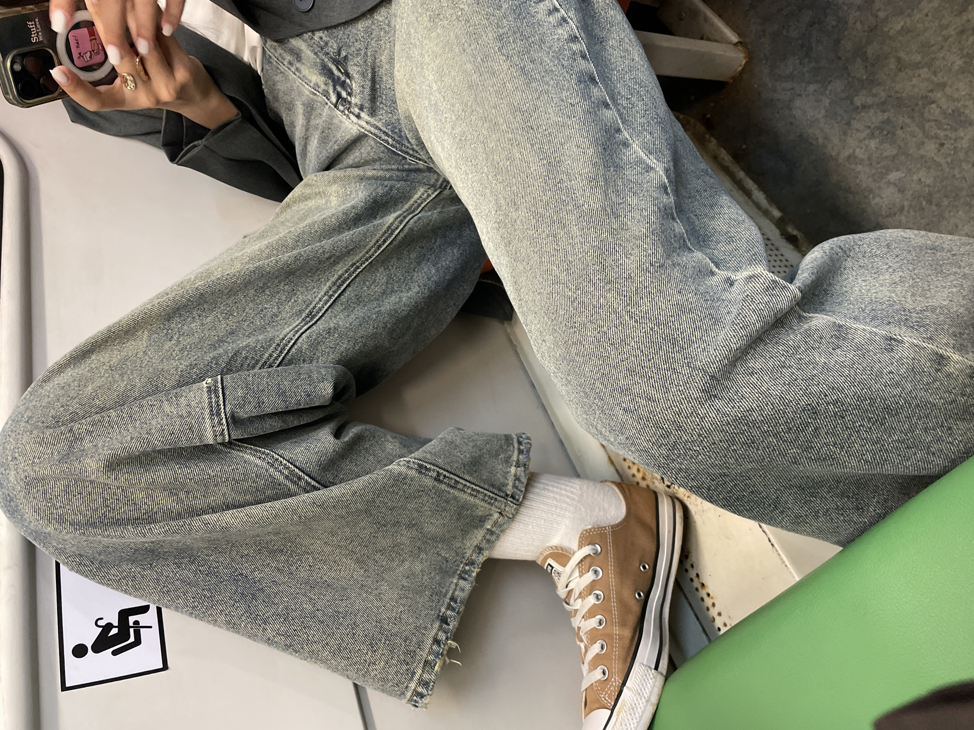 The image shows a person sitting with their legs crossed, wearing light blue wide-leg jeans, white socks, and brown Converse sneakers. The person is holding a cell phone in their left hand. The person is also wearing a gray blazer. A wheelchair accessibility symbol is visible on a white surface to the left. A green seat is also visible. The location is indeterminable without additional information.