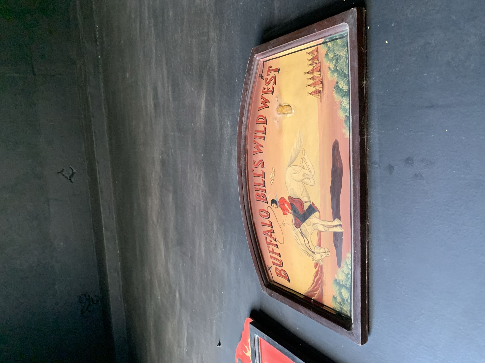 A framed, vintage sign depicting a Wild West scene hangs on a dark, textured wall. The sign is oriented vertically and is curved along the top edge. The artwork features a cowboy on horseback in the foreground, lassoing an unseen object. Behind him, a line of teepees is visible against a backdrop of rolling hills and a sunset or sunrise sky in shades of yellow, orange, and pink. Green trees are also depicted.
Visible text on the sign reads "BUFFALO BILL'S WILD WEST". There is a small, discolored patch on the sign near the text. The setting appears to be a wall, possibly in an indoor space, with a dark, somewhat rough texture. Another sign, partially visible at the bottom of the frame, appears to be red and black. The overall impression is one of historical or Western-themed decor.