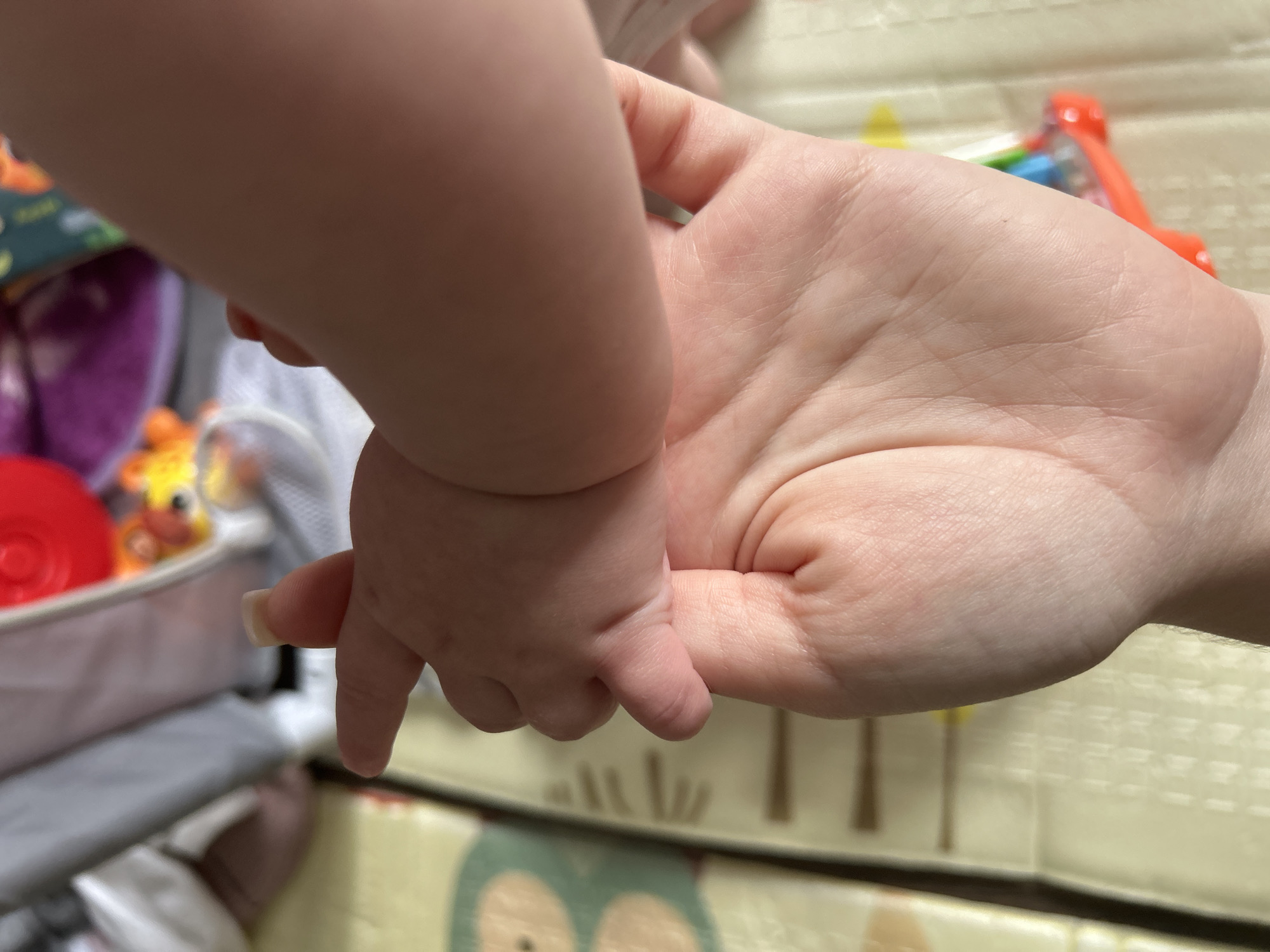 A close-up shot captures an adult's hand and a baby's hand interacting. The baby's hand, with its small fingers and a tiny fingernail, rests on the adult's palm. The adult's hand, showing visible lines and texture, cradles the baby's hand. The background is out of focus but appears to be a playmat with faint patterns and some blurred colorful objects, possibly toys. The lighting suggests an indoor setting.