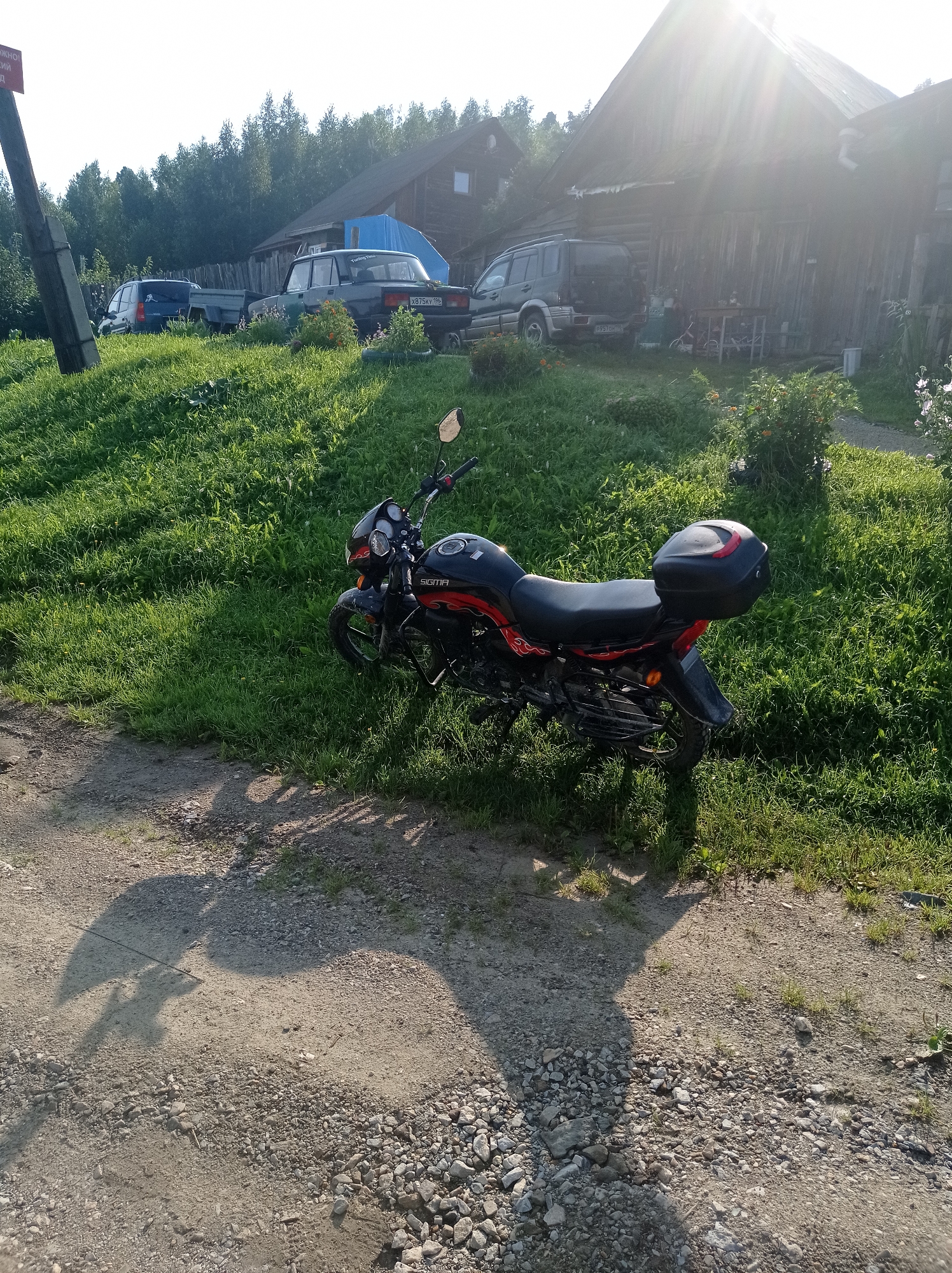 In an outdoor scene in Degtyarsk, Russia, a red and black motorcycle is parked on a grassy incline. The motorcycle features a black top case and red flame decals on its body. Behind the motorcycle, a weathered wooden house with a steeply pitched roof is visible, partially obscured by sunlight. In front of the house, several vehicles are parked, including a dark green SUV and a light-colored sedan. The ground is a mix of gravel and dirt. Trees and foliage surround the area, suggesting a rural or semi-rural setting. A shadow of what appears to be a person on a bicycle is cast on the ground in the foreground.