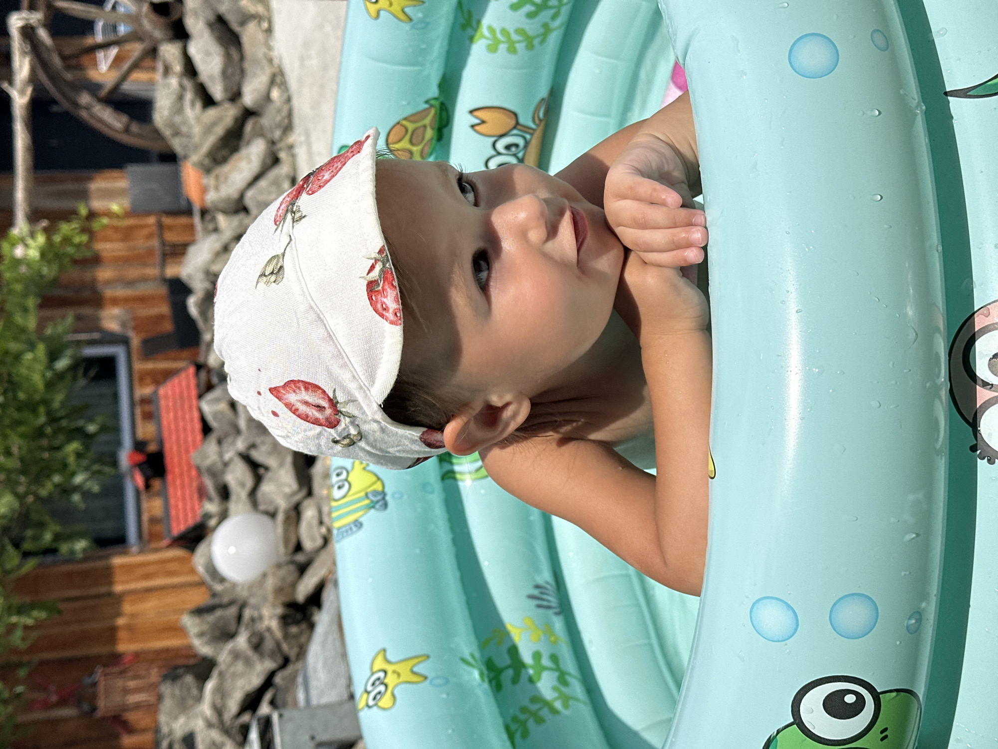 A baby is shown from the chest up, looking to the right with a hand near their mouth. The baby is wearing a white hat with a red strawberry pattern. They are in a light blue inflatable pool decorated with drawings of fish and seaweed. The pool is outdoors, with a background of wooden structures and stone. The lighting suggests it is daytime, possibly sunny. A round white light fixture is visible in the background. The scene appears to be in Wisła, Poland.