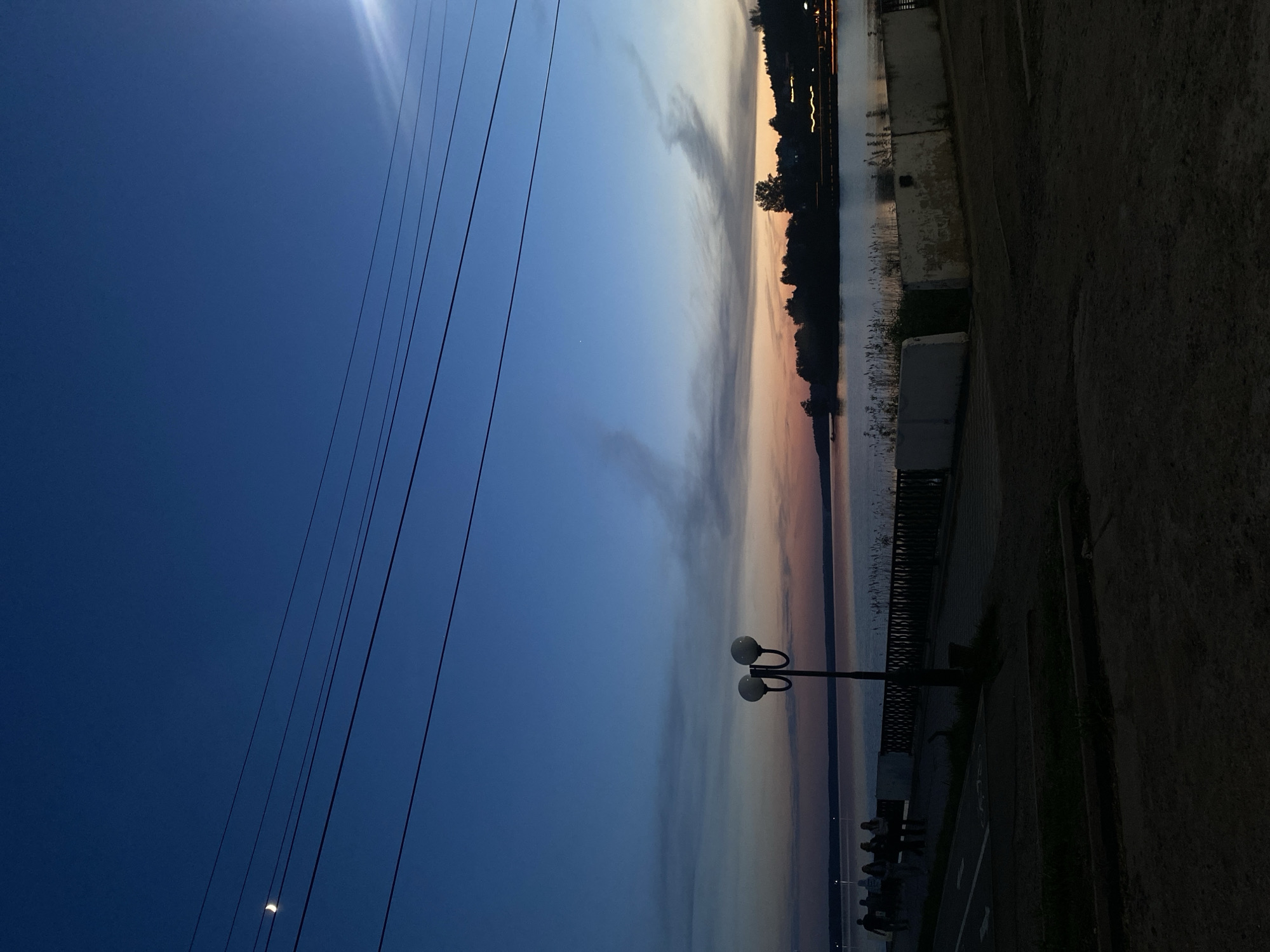 The image depicts a waterfront scene in Votkinsk, Russia, at dusk or dawn. A body of water reflects the pastel colors of the sky. A shoreline with trees and structures runs along the horizon. A paved walkway with a railing is visible along the water's edge. A distinctive lamppost with two spherical lights stands along the walkway. People are gathered in groups on the walkway. Overhead power lines cut across the upper left portion of the sky. The foreground on the right side of the image is dark.