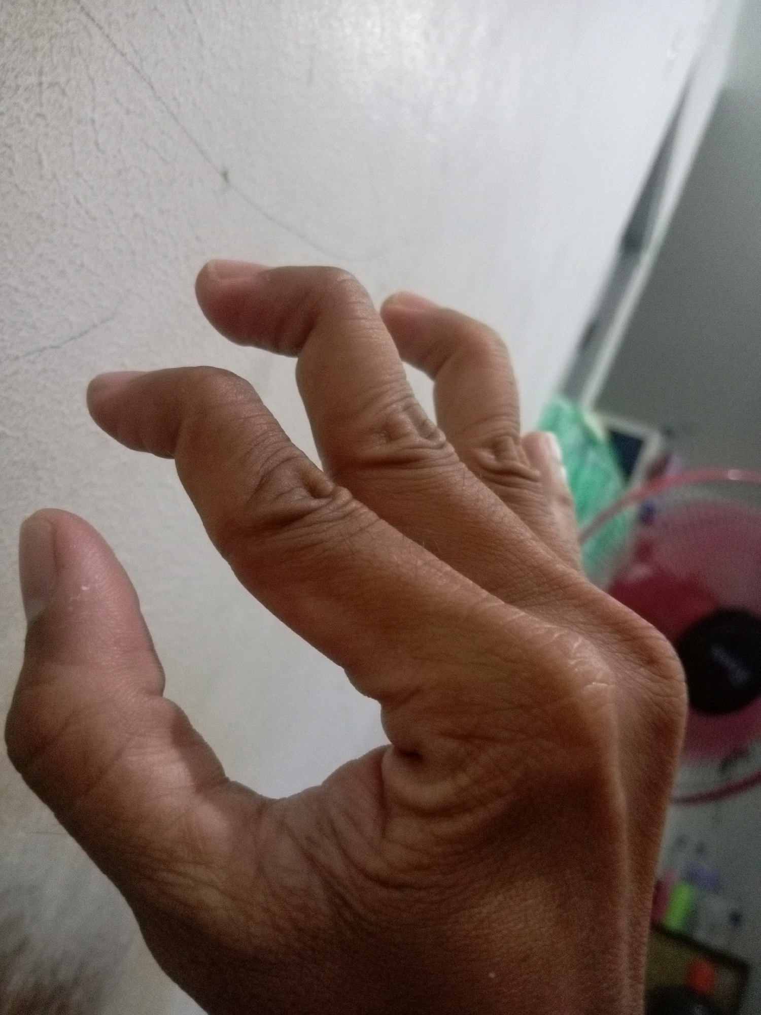 The image is a close-up, high-angle shot of a person's hand, likely taken indoors. The hand is positioned in front of a textured, light-colored wall that appears to be a plastered or painted surface with faint lines or cracks. The skin of the hand is tanned, and the fingers are bent in a claw-like or grasping position. Some of the finger joints appear prominent. The background is out of focus but suggests an indoor environment with hints of other objects, including what looks like part of a pink fan and possibly some green and blue material. There are no clear indications of the specific location within Batam, Indonesia, beyond it being an interior space. The lighting is somewhat diffuse, casting soft shadows on the hand.