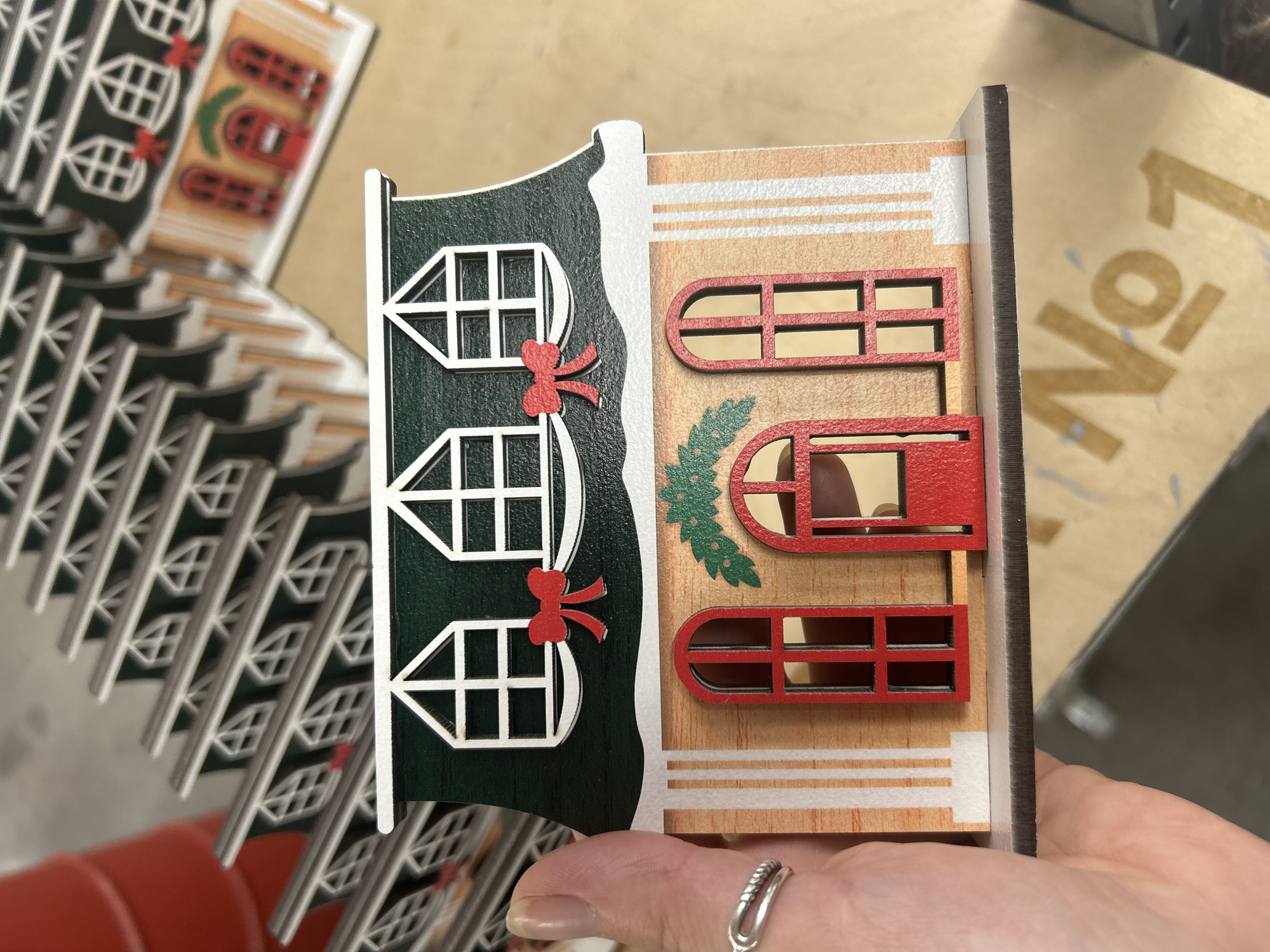 A hand with a silver ring on the index finger is holding a miniature wooden Christmas house. The house is decorated with green roofing, white windows with red bows, and a light brown facade with white vertical stripes and red arched doors and windows. In the background, rows of similar miniature houses are stacked, creating a festive display. The environment appears to be indoors, possibly a store or workshop, with a wooden surface visible beneath the houses. There is no discernible information in the image that can be used to determine the location in the city of Ternopil, Ukraine, or any specific actions or interactions occurring.