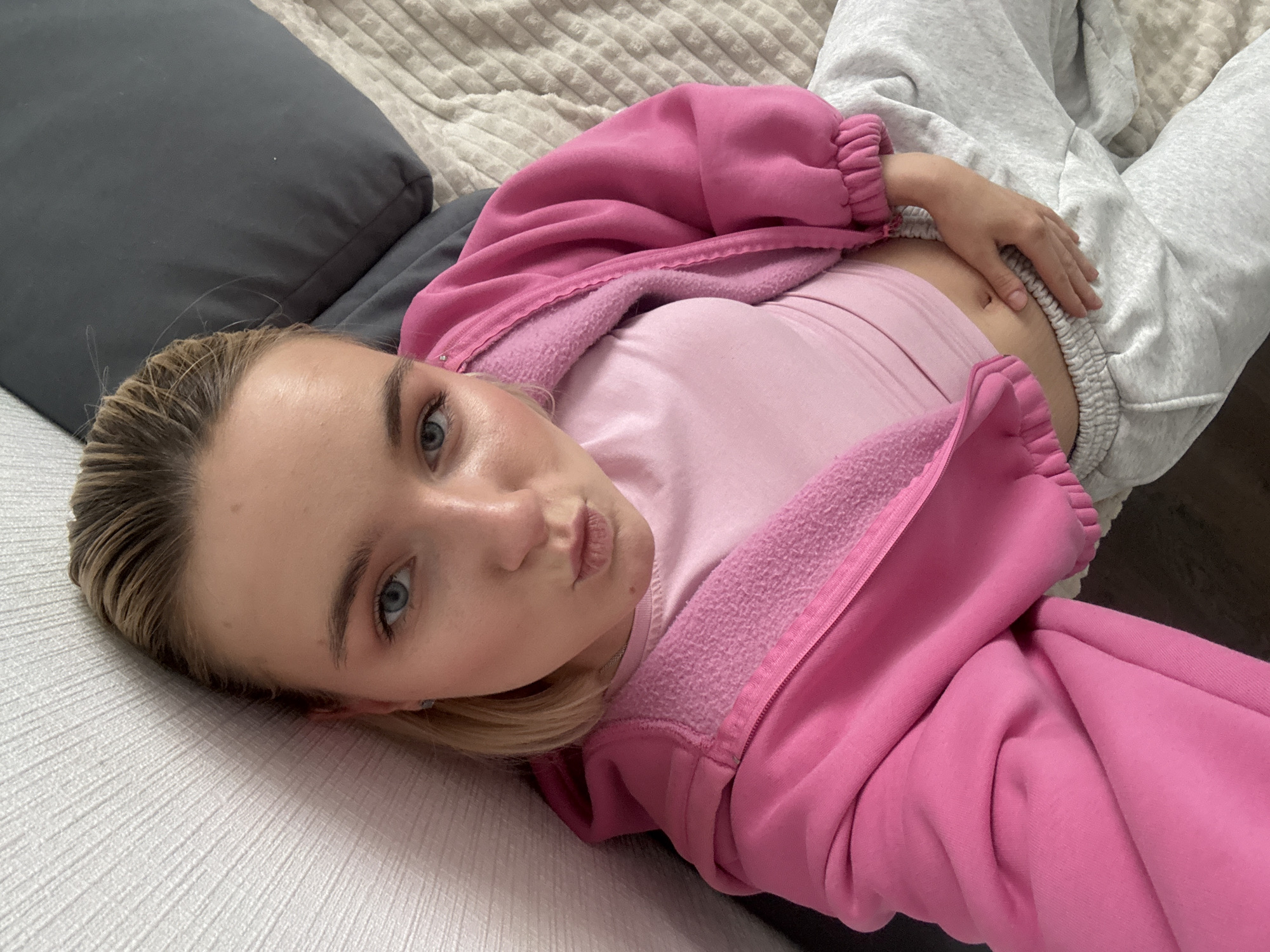 A young woman is lying on a light grey and white patterned surface, possibly a bed or sofa. She is wearing a light pink t-shirt, a bright pink zip-up hoodie, and grey sweatpants. Her blonde hair is pulled back, and she is looking directly at the camera with a slightly pouting expression. Her right hand rests on her stomach. The background includes a dark grey pillow and part of the bed covering. The photo seems to be taken in a private indoor setting. There is no information to suggest that this image was taken in the city of Vsevolozhsk, Russia.