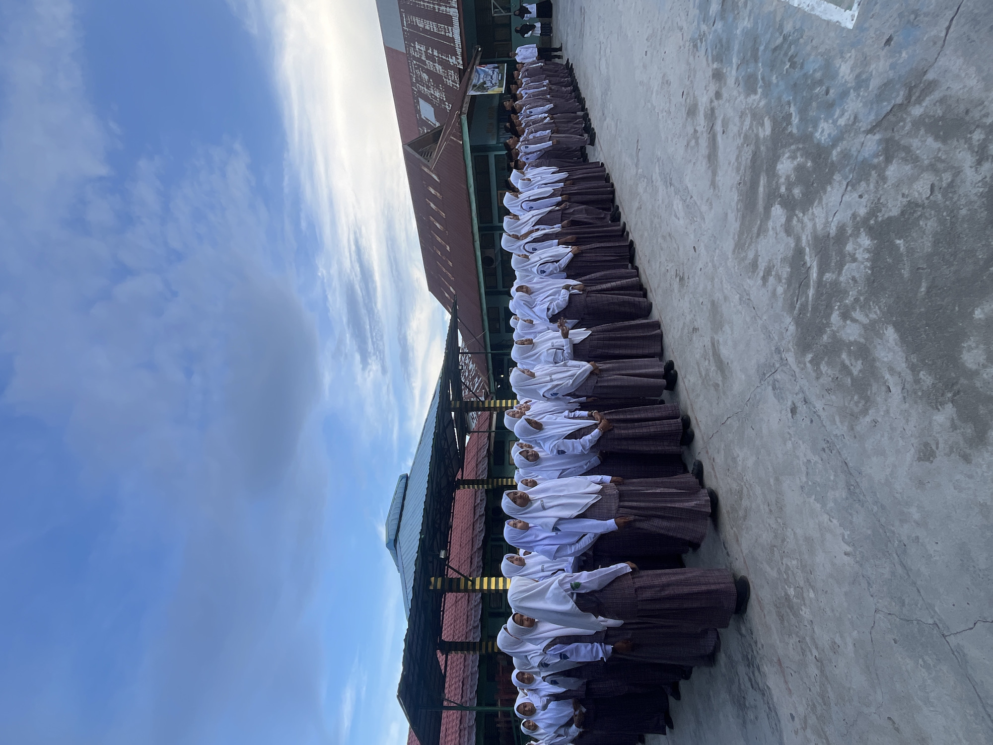 In Lhokseumawe, Indonesia, a large group of students, predominantly girls wearing white hijabs and brown plaid skirts, stand in formation on a concrete outdoor area. Some of the girls are on the left side of the image, facing towards the right. In the center and right portions of the image, a mixed group of students, including boys in uniforms, are also standing in formation, facing towards the left. Behind the students, there are buildings with red and green roofs, and a covered outdoor area with a dark blue roof supported by yellow poles. The sky above is partly cloudy with a mix of blue and grey. The scene appears to be an outdoor school assembly or event.