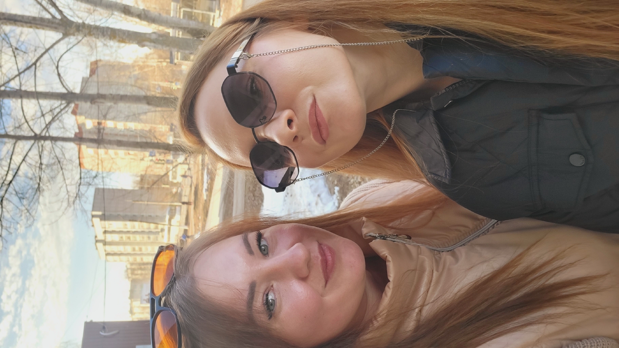 Two women are posing for a selfie outdoors in Chaikovsky, Russia. The woman on top is wearing sunglasses with a chain and a black jacket. The woman on the bottom is wearing sunglasses and a beige jacket. Bare trees and apartment buildings are in the background.
