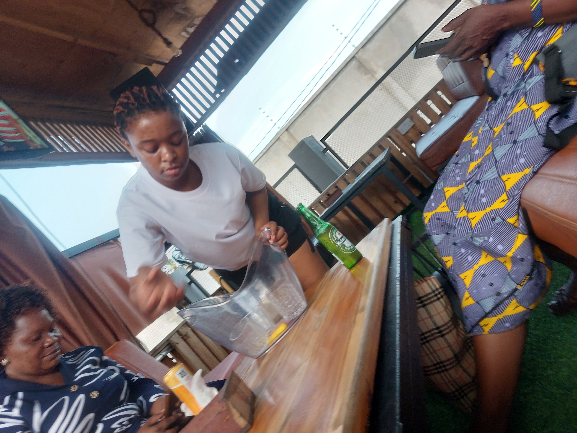 Here is a description of the image:
The image is taken at an outdoor establishment, possibly a restaurant or bar, in Dar es-Salaam, Tanzania. A young woman in a white t-shirt is preparing a drink using a plastic pitcher on a wooden bar. There is a green beer bottle next to her on the bar. Two patrons are visible; one is sitting to the left of the frame, wearing a patterned top. Another patron in a blue and yellow patterned dress sits on a leather seat, looking at a mobile phone. A checkered handbag rests near their feet. The area is covered by a roof constructed of wooden slats.