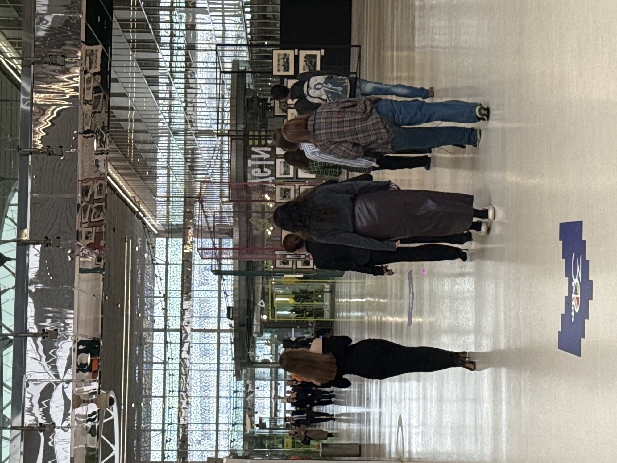 The image depicts an indoor scene within a large, modern building, likely an art gallery or exhibition hall. The perspective is from behind a group of people, who appear to be walking through or observing an exhibition.
In the foreground, a person with long, dark brown hair is visible from the back, wearing black attire, suggesting a sleek, contemporary style. Behind them, several other individuals are also seen from the back, forming a loose queue or moving through the space. One person wears a plaid shirt and blue jeans, while another further back has a dark jacket, blue jeans, and a backpack featuring a white graphic design.
The environment is characterized by significant natural light, indicating daytime. The left side of the frame features highly reflective, silver-colored architectural elements that show distorted reflections of the surroundings, including parts of the ceiling and other structures. The background reveals a vast, grid-like structure, possibly a large skylight or window system, allowing ample light to illuminate the space.
Throughout the middle ground, several display stands are visible, holding numerous framed pictures, likely photographs or artworks, suggesting an art exhibition is taking place. On one of these display elements, partially visible Russian text reads "сети," which translates to "networks" or "nets."
The floor is light-colored and somewhat reflective, capturing shadows and reflections of the people and light sources. On the right side of the floor, a prominent blue graphic is visible, displaying "30 P" (where 'P' likely denotes rubles or years) and a stylized circular logo containing the letters "C" and "P". The overall atmosphere is bright and open, indicative of a public cultural venue.