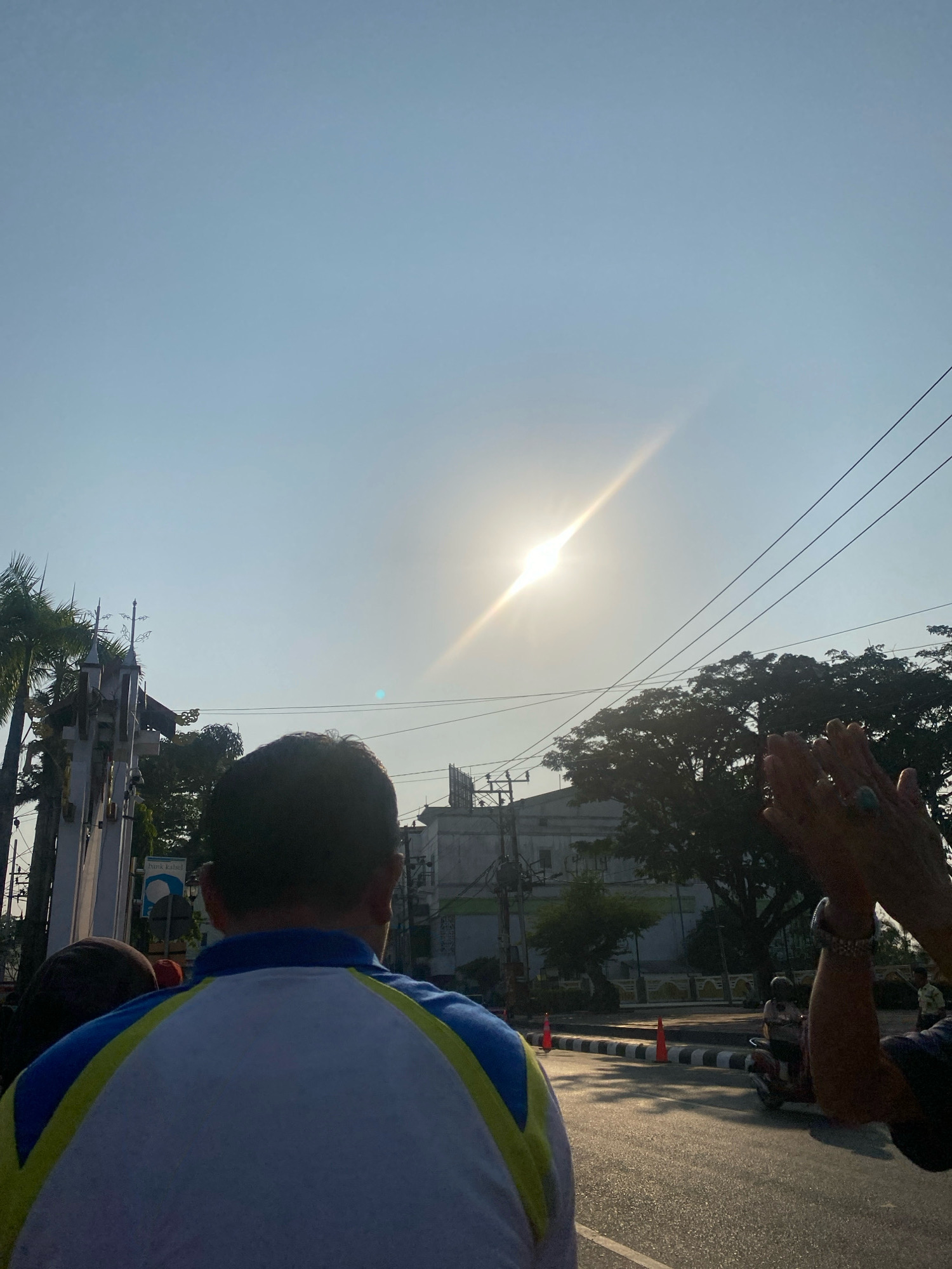 In Banjarmasin, Indonesia, on a clear day with the sun shining brightly in the sky, a group of people are gathered outdoors on a street. The perspective is from behind a person wearing a blue, yellow, and white shirt, who is looking towards the sun. In the foreground, partially obscuring the view, are the backs of several individuals. To the left, a tall, ornate structure, possibly a monument or part of a building, is visible with palm trees nearby. Further back, the silhouette of leafy trees frames the scene. A road runs through the middle ground, with traffic cones and a scooter with a rider suggesting a public street or event. On the right, a pair of hands are raised, with jewelry on the wrist, appearing to clap or gesture. The overall atmosphere suggests an outdoor gathering or event, possibly a public celebration or observation, under the bright Indonesian sun.