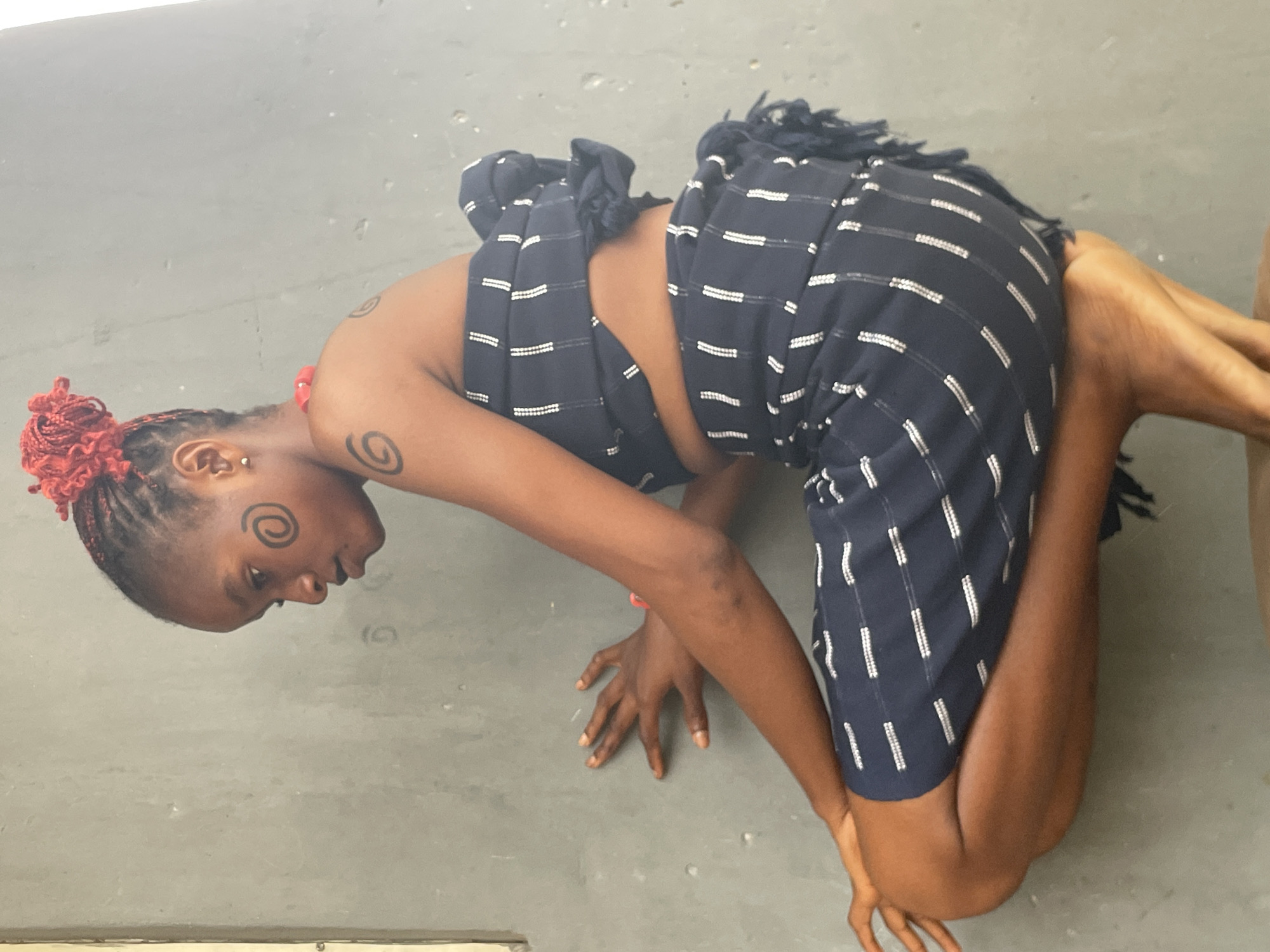 An image captured in Badagry, Nigeria, features a person with dark skin in a low crouching or kneeling pose on a plain, light gray floor. The individual's body is bent forward, with their head tilted downwards. Both hands are placed on the ground in front of them, fingers spread, supporting their weight. They are dressed in a dark blue fabric with white dashed stripe patterns, resembling a traditional wrapper or garment, which features fringed edges. The person has red braided hair styled upwards on their head. Visible accessories include a small silver ear piercing on the left earlobe and a reddish band on the right wrist. Distinctive black spiral markings are present on the left cheekbone and on both upper arms. The lighting is bright and uniform. The person's expression is neutral, indicative of focus on maintaining the physical pose. No other objects or specific environmental details are visible in the background.