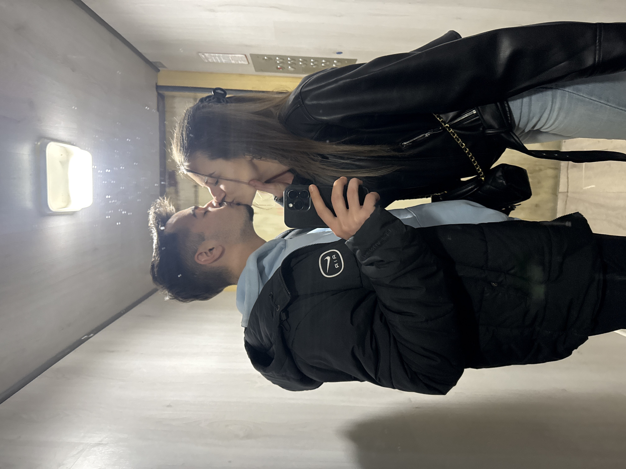 A woman in glasses and a black leather jacket is kissing a man in a black puffer jacket inside what appears to be an elevator. The woman is holding a smartphone and seems to be taking a selfie. The elevator is white with a square light fixture on the wall. There are also what seem to be bubbles in the air. The location might be in Sofia, Bulgaria.