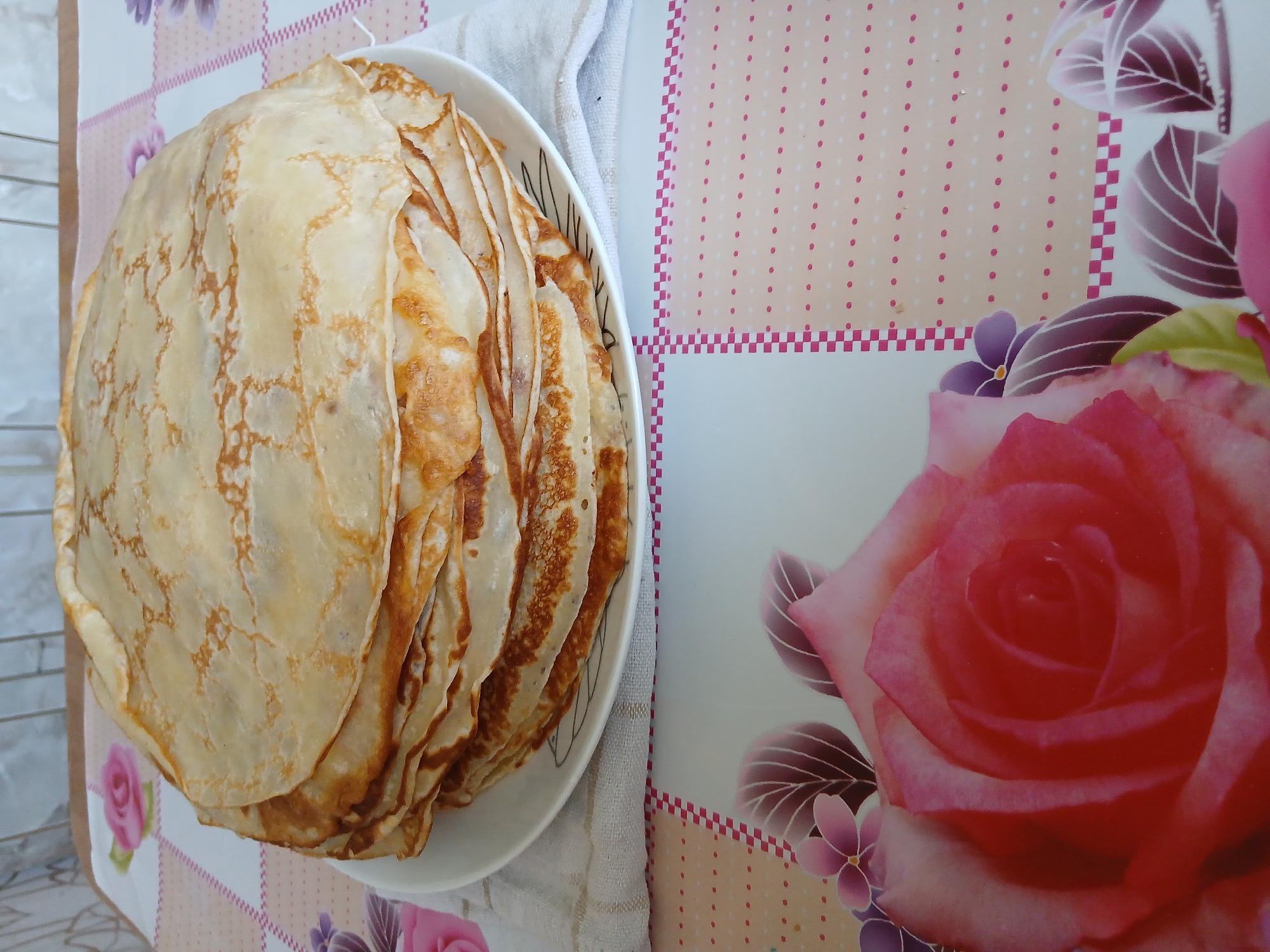 A stack of thin, golden-brown crepes sits on a white plate, which is resting on a white tea towel. The crepes have irregular edges and some are folded. The plate and crepes are on a table covered with a tablecloth. The tablecloth has a checkered pattern along the top and a floral pattern at the bottom, featuring a large pink rose and purple flowers. The background shows a tiled wall. The scene appears to be indoors. There are no people visible in the image. The location is not specified in the image or its context.