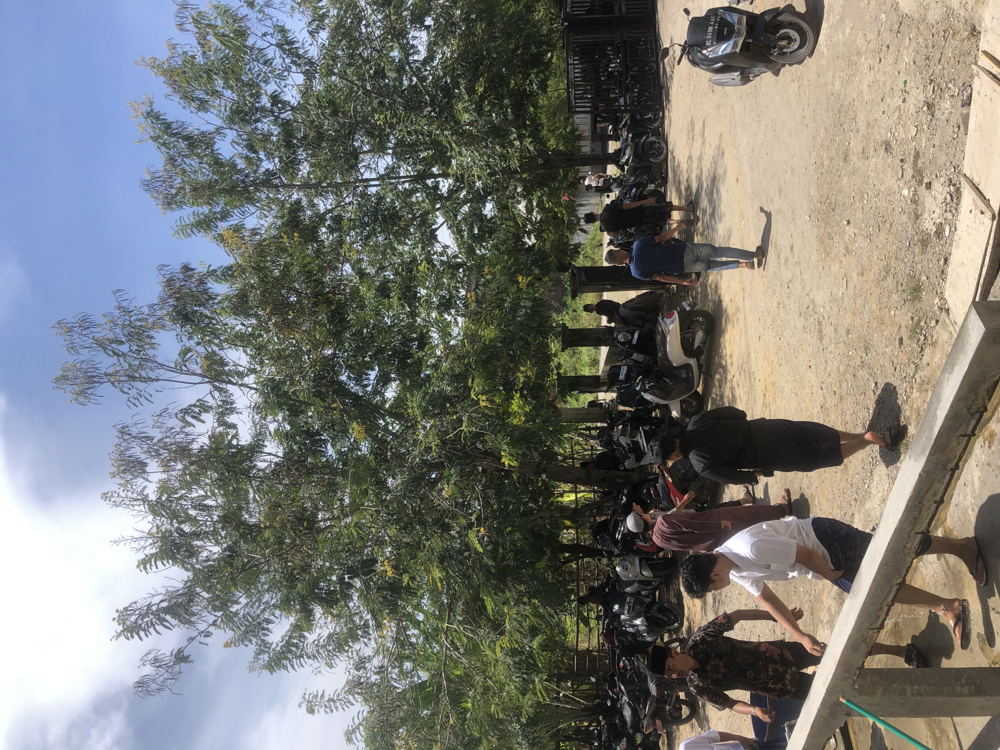 In Lhokseumawe, Indonesia, on a bright, sunny day, a group of men are gathered outdoors. The scene is characterized by a dusty, unpaved ground and a large number of parked motorcycles. Several trees with dense green foliage dominate the upper portion of the image, casting shadows.
In the foreground, a man in a white shirt and dark shorts is leaning over a concrete structure, with water splashing around his feet. Another man to his left, wearing a patterned shirt and a black peci (skullcap), is holding a blue bucket. Further back, a man in a dark robe walks away from the camera. To the right of the frame, a man in a blue t-shirt and jeans walks towards the background. Other individuals are visible among the parked motorcycles, some appearing to be arriving or departing. The motorcycles are of various models and colors, predominantly dark. In the distance, a black fence and more buildings are visible. The sky is partly cloudy with a clear blue sky visible in patches.