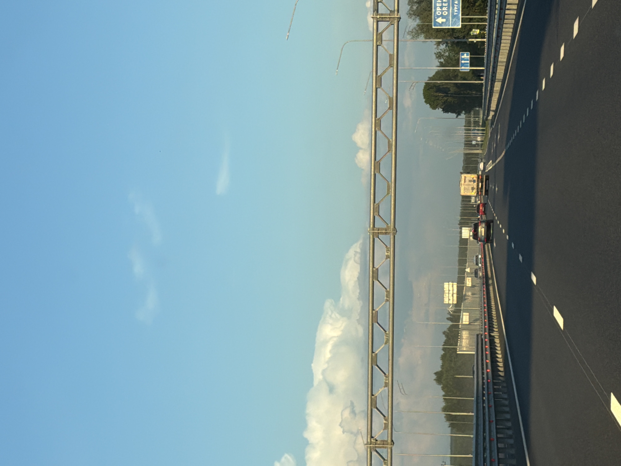 The image depicts a highway scene in Zimnyaya Gorka, Russia, under a clear blue sky with scattered clouds. The perspective is from a moving vehicle, looking forward. In the foreground, a dark asphalt road with white lane markings is visible. A metal guardrail lines the right side of the road.
In the middle ground, several vehicles are traveling on the highway, including cars and a truck. A large overhead gantry structure with metallic supports spans across the highway. Streetlights are positioned along the roadside. In the distance, a dense line of trees forms the horizon.
On the right side of the highway, a blue directional sign with white text and symbols is prominently displayed. The text on the sign appears to read "ОРЕНБУРГ" (Orenburg) and "ТУРГАЙ" (Turgay), indicating directions to these locations. Other smaller signs are also visible in the distance. The overall environment suggests a typical highway setting outside of a populated area, likely during daytime.