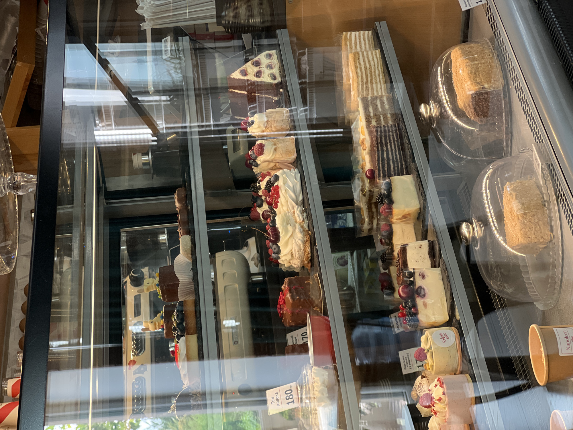 This is a medium shot photograph of a refrigerated display case filled with various cakes and pastries. The display case is made of glass and metal, with multiple shelves. The lighting appears to be from within the case and from overhead.
On the top shelf, there are several small cakes decorated with chocolate and fruit, including two with white bunny figures on top. Below this, on the second shelf, there are several more elaborate cakes, including one with meringue and berries, and a triangular slice of cake with cream and cherries.
The third shelf features a selection of smaller individual cakes and pastries, some with fruit toppings. A label reads "Три молока 180грн". Another label is partially visible, reading "170".
The bottom shelf displays several multi-layered cakes, including what appears to be a "Napoleon" cake. Two of these cakes are covered with glass domes. A brown paper cup with a white label is also visible on this shelf.
The background shows reflections in the glass of the display case, including what appears to be trees and sky, suggesting an outdoor environment visible through a window. The overall impression is of a well-stocked bakery or patisserie. The provided information indicates the location is Fontanka, Ukraine.