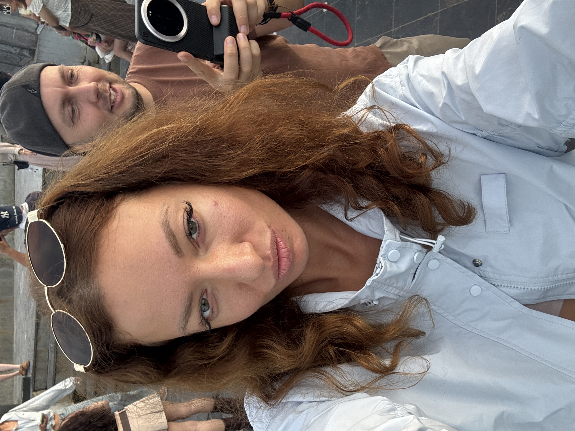 A woman with auburn hair is facing the camera, wearing sunglasses on her head and a light blue jacket, seemingly taking a selfie with a man in the background wearing a gray cap and holding a black camera with a red strap. The setting appears to be Lũng Cú Commune, Vietnam, indicated by the background which seems to be a tourist location given the other people present and the overall setting.