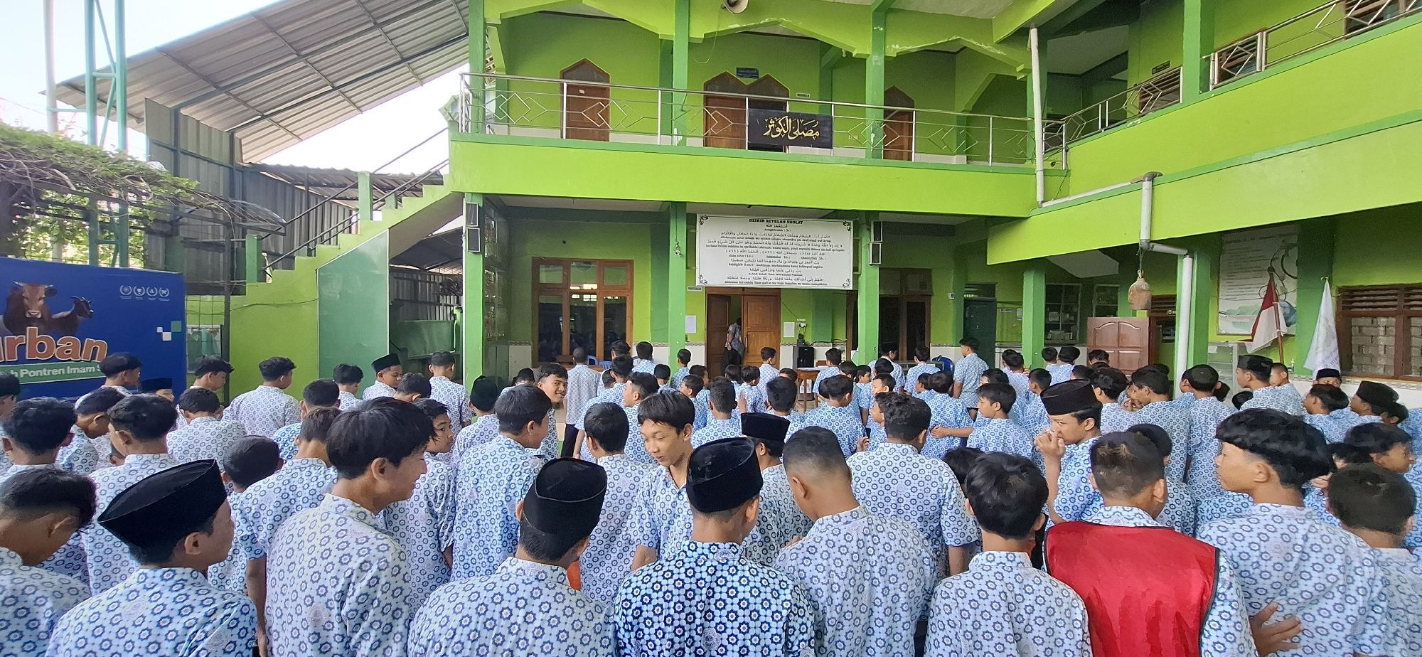 In Wonorejo, Indonesia, a group of young men, likely students at a religious boarding school (pondok pesantren), are gathered in an open courtyard. They are all dressed in blue and white patterned shirts and many are wearing black peci caps. The architecture of the building is predominantly bright green, with multiple levels and balconies visible. A large banner with Indonesian text and images of cattle is displayed on the left side of the frame, suggesting a connection to agricultural activities or a sponsored event. A whiteboard or poster with religious text in Arabic is mounted on a bright green wall. The scene appears to be a communal gathering, possibly for prayer, an assembly, or an educational session, under the bright daylight. The overall environment suggests a structured, educational, and community-oriented setting within a tropical climate.