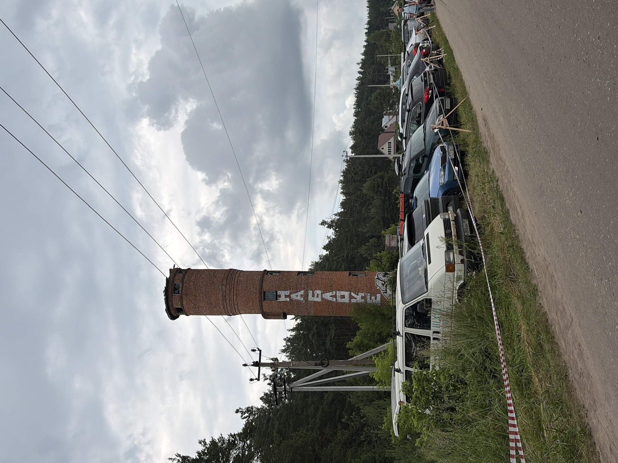 A brick water tower with Cyrillic lettering "НАБАБКЕ" spray-painted vertically on its side is visible in Il'inskiy Pogost, Russia. The tower is partially obscured by trees and power lines. In the foreground, a row of parked cars is lined up along a dirt road, with a white van positioned closest to the camera. Red and white caution tape is stretched across the grassy area in front of the van. The sky is overcast, with large, grey clouds dominating the upper portion of the image. The environment appears to be a semi-rural or industrial area with dense forestation in the background.