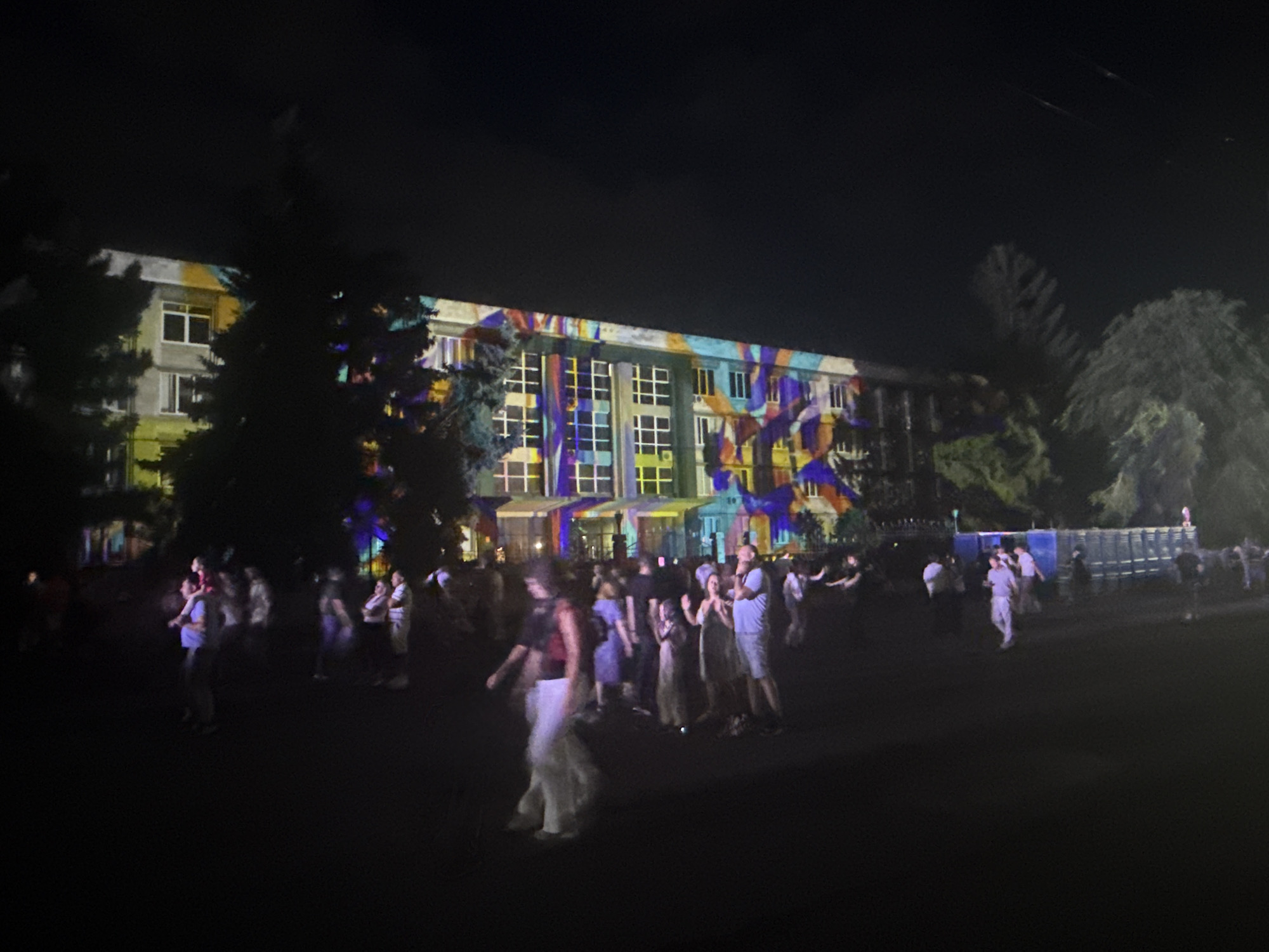 In Chișinău, Moldova, a nighttime scene unfolds in front of a building illuminated by a vibrant light projection. The building, with multiple windows arranged in a grid pattern, is adorned with abstract, colorful shapes in shades of blue, yellow, purple, and red. Dark trees with dense foliage are silhouetted against the illuminated facade. A crowd of people is gathered in the foreground and mid-ground, some appearing to be walking, others standing and conversing. Their faces are not clearly discernible due to the low light conditions and the distance. The ground appears to be a dark, unpaved area, possibly a public square or open space. In the background, a large, dark structure, possibly a fence or a series of containers, is visible. The overall atmosphere suggests an outdoor event or festival.