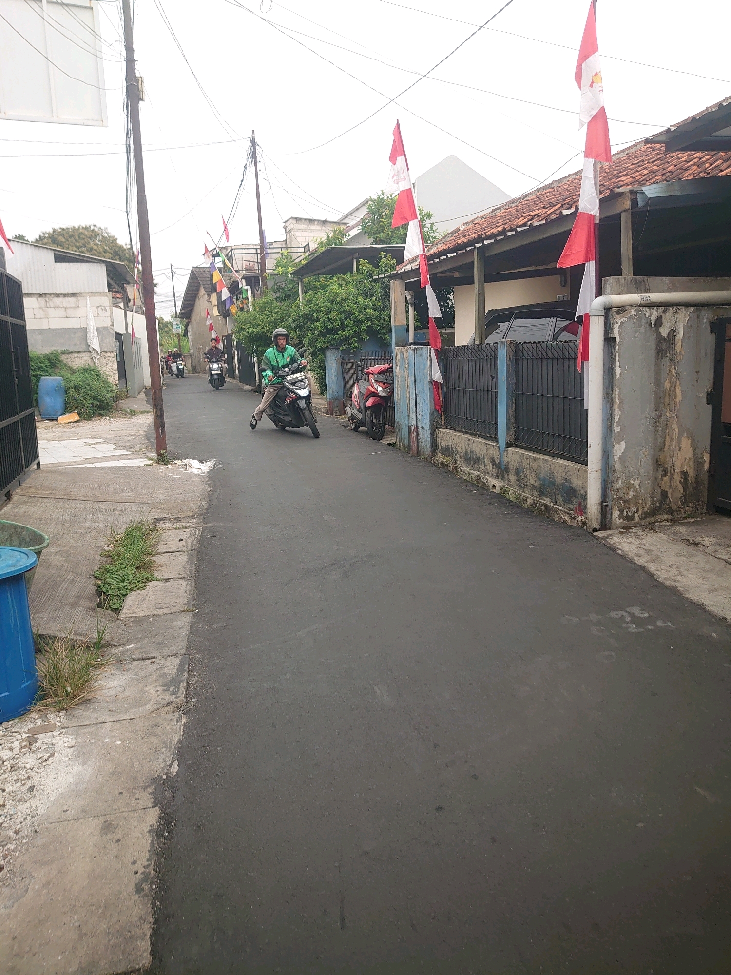 The image depicts a narrow, asphalt-paved street in Depok, Indonesia, lined with residential buildings and adorned with numerous red and white Indonesian flags. A person wearing a green jacket and helmet is riding a motorcycle towards the foreground, leaning to the left as they navigate the street. Several other motorcycles are visible further down the road, also traveling in the same direction. On the right side of the street, a weathered concrete wall with a black metal fence runs along the property line, separating the road from a house with a tiled roof. Red and white flags are also attached to this fence. The left side of the street features a concrete sidewalk, a blue trash bin, and some overgrown vegetation. The sky is overcast, indicating a cloudy day. The overall scene suggests a neighborhood decorated for a national celebration.