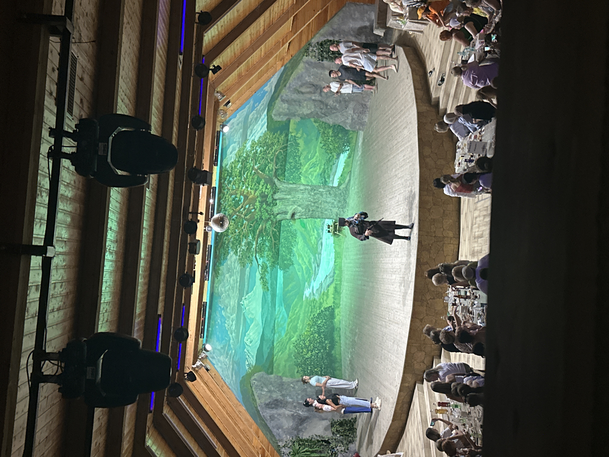 An audience of approximately 40 people is seated at tables arranged on a lower level, observing a performance on a raised, circular stage. The stage backdrop depicts a lush green landscape with mountains, a river, and a large tree. On the stage, a single performer in traditional dark clothing and a hat stands center, while a group of five individuals, three women and two men, are positioned on the left side of the stage, and another group of five men stand on the right. The venue appears to be an open-air amphitheater with a wooden, sloped roof structure. Lighting fixtures are visible suspended from the ceiling, and a disco ball hangs above the stage. The overall setting suggests a cultural event or performance taking place in Bolshoy Kichmai, Russia.
