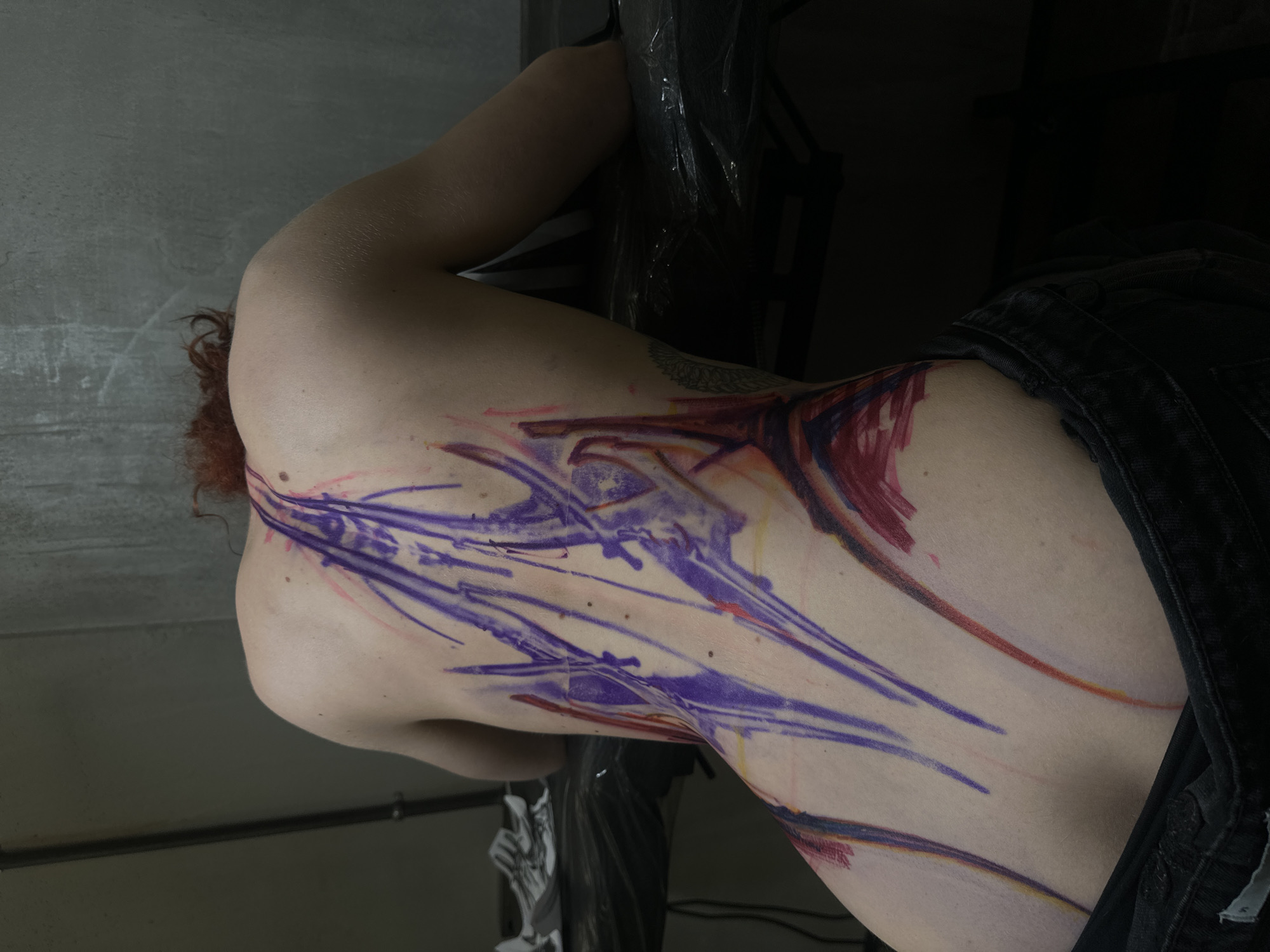A person is lying prone on a surface, with a large tattoo spanning their back and side. The tattoo design features strokes of purple and red. The person has fair skin and reddish-brown hair. They are wearing dark jeans. The environment appears to be an indoor space, with gray walls and a dark area in the background containing what looks like equipment covered in plastic. There is no indication of the location being in Moscow, Russia.