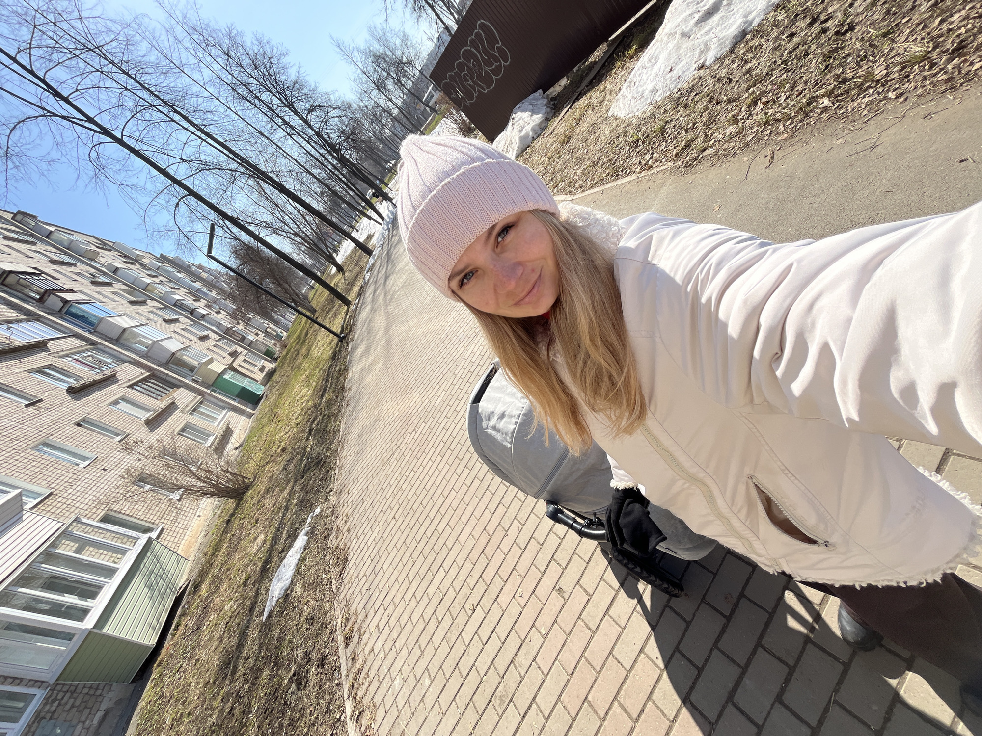 A young woman is taking a selfie on a brick sidewalk. She is wearing a light pink beanie and a white jacket with a beige trim. She is holding a grey baby stroller. In the background, there is a multi-story brick building with many windows, a line of bare trees, and patches of snow on the ground. A brown fence with graffiti is also visible. The location is Chaikovsky, Russia.