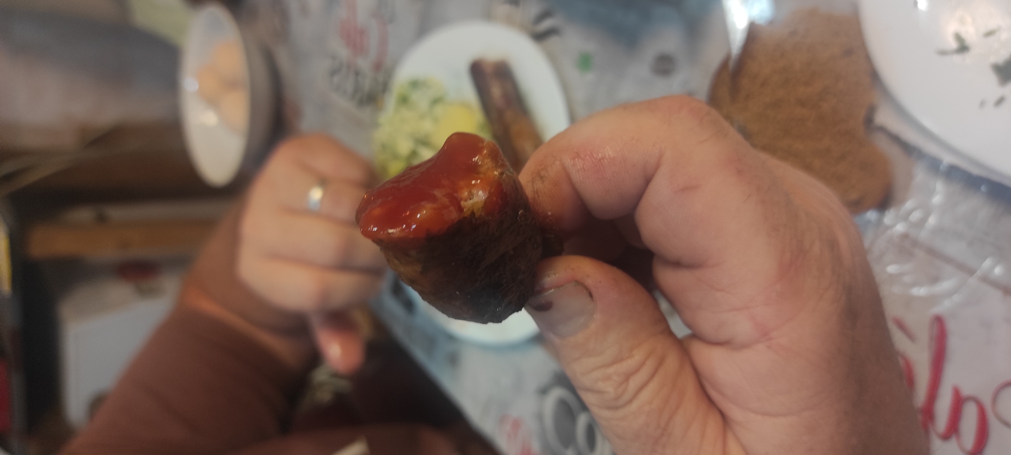 In Kobeliachok, Ukraine, a close-up image shows a person's hand holding a piece of food, possibly a sausage, covered in ketchup. The food item is dark brown and appears to be textured, with a shiny red ketchup glaze. The hand holding the food has visible fingernails and skin texture. In the background, slightly out of focus, is a white plate with what appears to be a side dish of mashed potatoes or similar and another sausage. A white bowl with round, light-colored objects, possibly pastries or dumplings, is also visible. The setting suggests a meal or gathering, with a patterned tablecloth and glimpses of furniture.