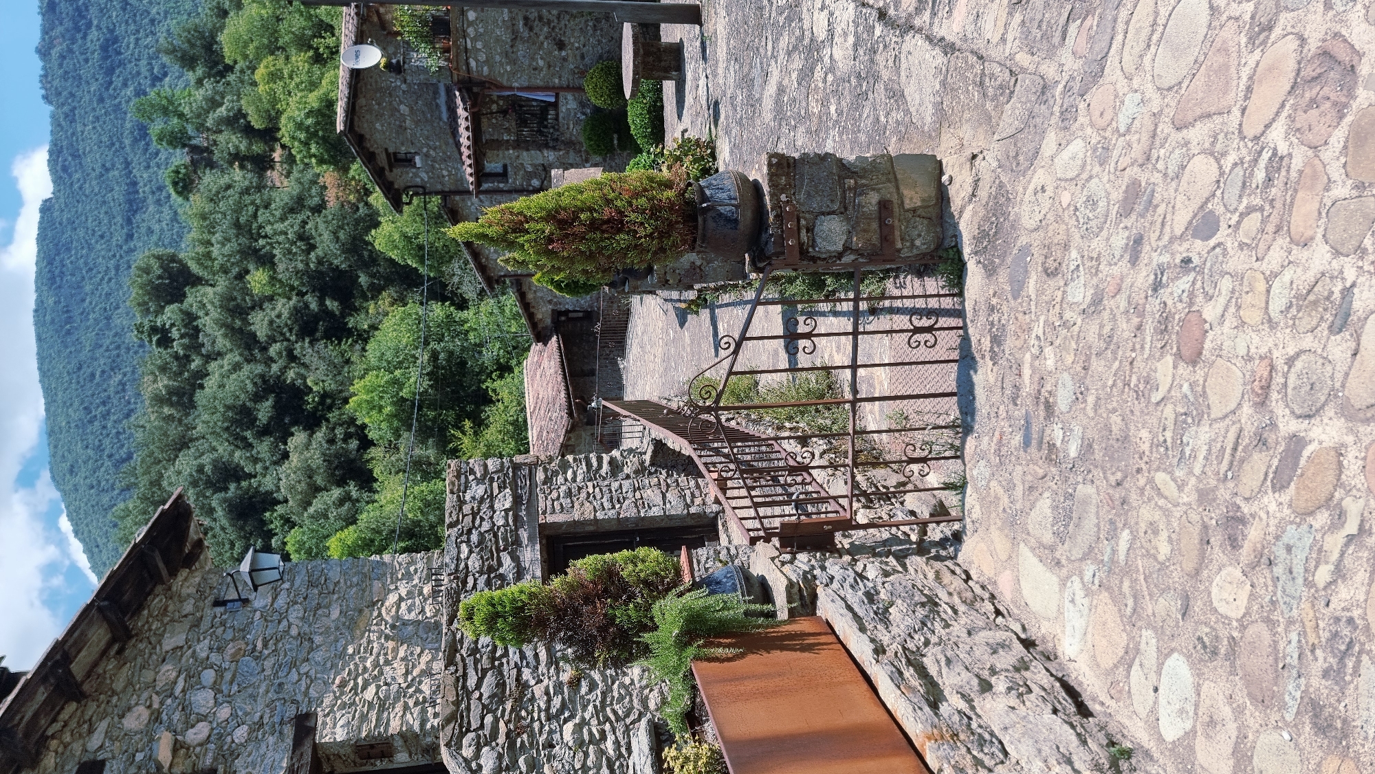 The image depicts a rustic stone pathway in what appears to be a rural village, possibly in the mountainous region near Camprodon, Spain. The pathway is paved with irregularly shaped, light-colored stones, interspersed with lighter grout. To the left, a stone building with a dark wooden door and a wall constructed of varied grey stones dominates the foreground. A rusty metal planter box sits at the base of this building. Two potted evergreen trees, one dark green and the other with reddish-brown tips, are placed strategically along the pathway. A decorative, rusty metal gate with swirling patterns stands open, leading further into the scene. Beyond the gate, the pathway curves to the right, flanked by similar stone buildings. One building in the mid-ground features a red-tiled roof and a satellite dish on its exterior wall. In the background, a dense forest of lush green trees covers rolling hills, culminating in a large, verdant mountain under a bright blue sky with scattered white clouds. The overall atmosphere is one of tranquil, old-world charm. No people are visible in the image, and no actions or interactions are depicted.