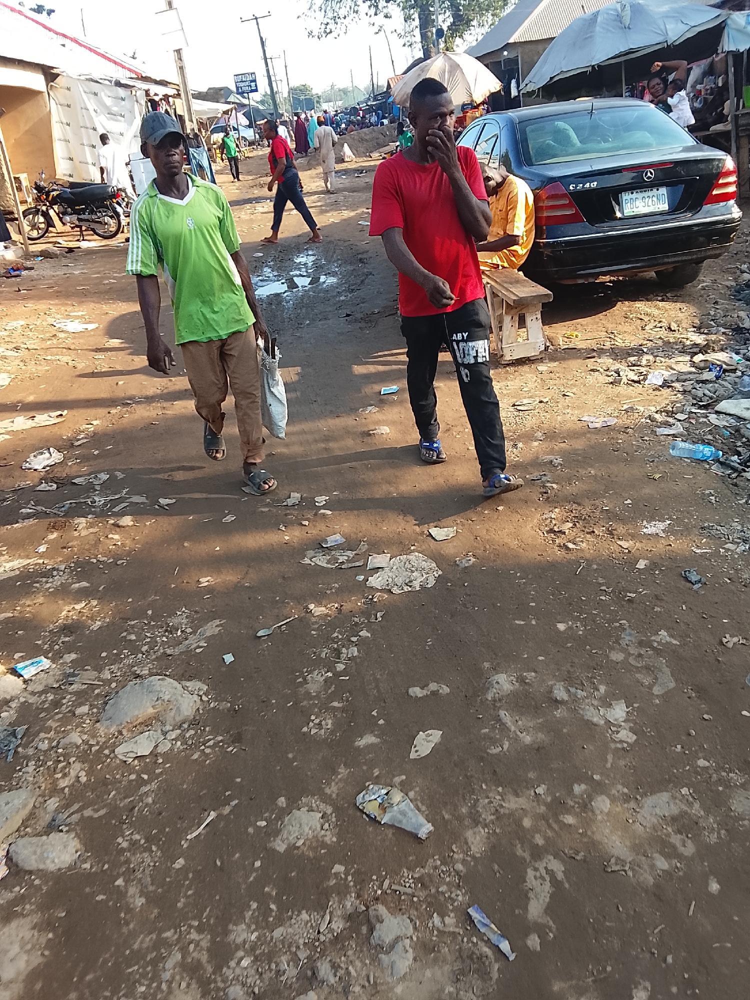 The image depicts a street in what appears to be a market area in Abuja, Nigeria. The ground is dirt and gravel, littered with trash. Two men are the main subjects: one wears a green shirt and khaki pants, carrying a bag; the other wears a red shirt and black pants with white lettering, covering his mouth with his hand. A black Mercedes C240 is parked on the right side of the frame, with the license plate RBC-926ND visible. Other people, motorcycles, and market stalls are in the background.