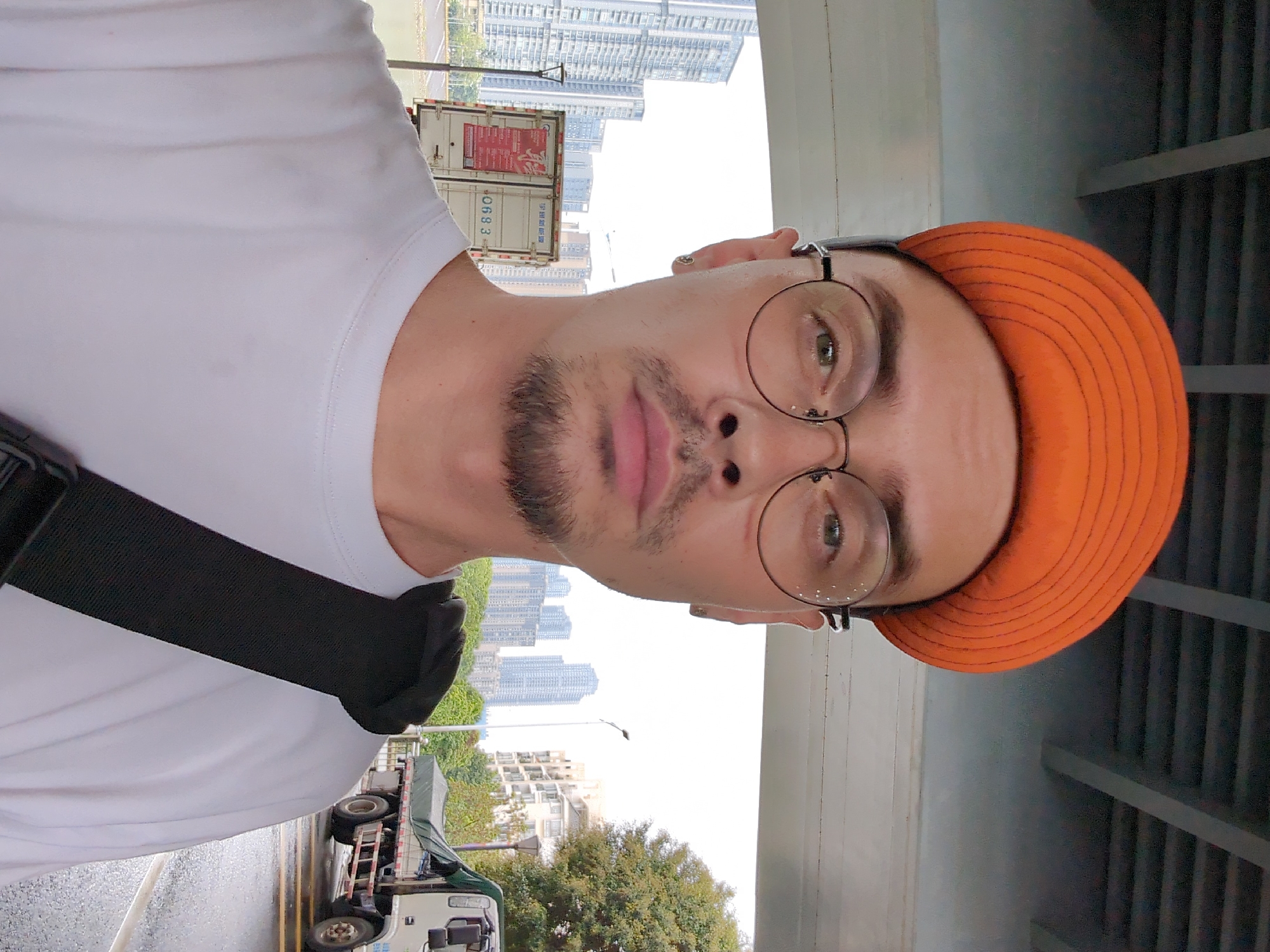A selfie photograph shows a person wearing a white t-shirt, an orange hat, and wire-rimmed glasses. The person has light skin, short brown hair, and a patchy beard. A black strap is visible across their shoulder. The background shows a cityscape, with tall buildings visible behind trees and other structures. A truck is parked on the street. The photo appears to be taken under a bridge or elevated structure, as indicated by the visible beams. The location is Shunde District, China, as inferred from the architecture and city features.