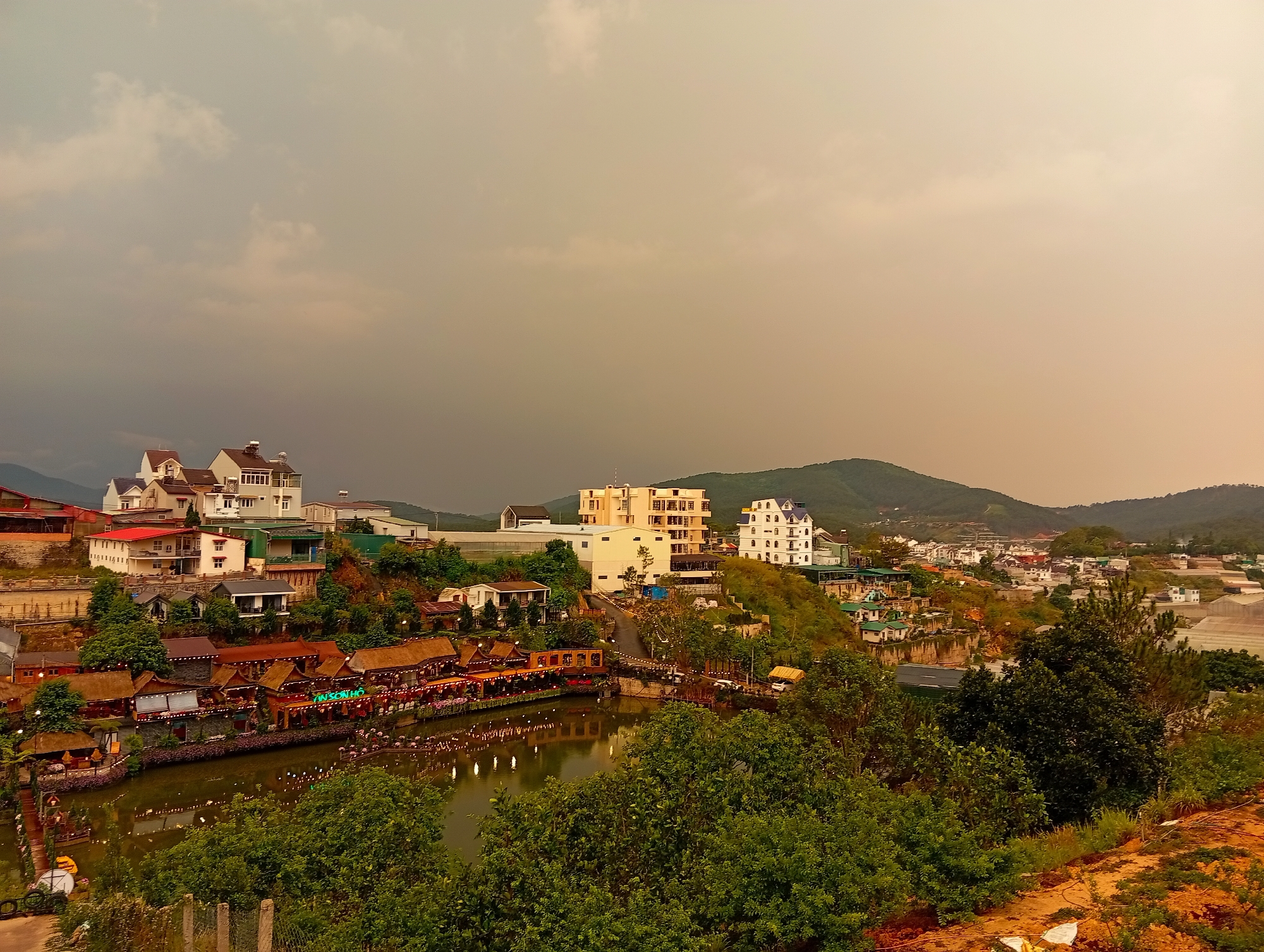 The image depicts a scenic view of a residential and commercial area in Da Lat, Vietnam. In the foreground, a body of water reflects light, surrounded by lush green trees and vegetation. A row of buildings with traditional thatched roofs lines the water's edge, suggesting restaurants or shops. These structures are adorned with lights, adding to the atmosphere.
Behind the water, a hillside is dotted with various buildings, ranging in size and architectural style. A few multi-story buildings stand out against the backdrop of the sky. Further back, rolling hills are visible in the distance under an overcast sky, which is shaded in hues of orange and grey. A road winds through the buildings, connecting the different levels of the landscape. The general setting suggests a densely populated area built on a hillside overlooking a body of water.