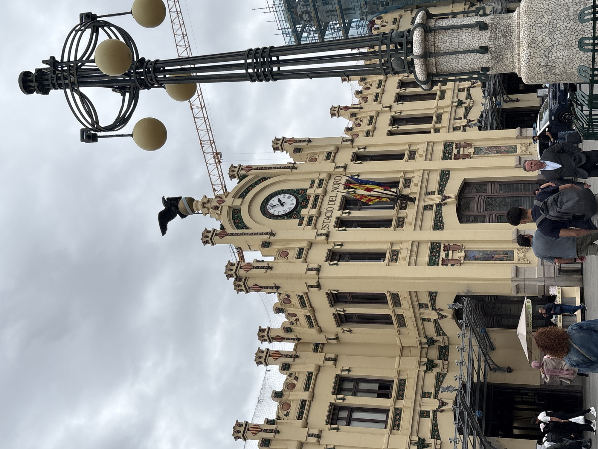 The Estació del Nord is located in València, Spain. The train station building is constructed of tan stone, with mosaics and ornate decoration. A lamp post with 5 spherical lights is on the left side of the image, and parts of a crane and scaffolding are visible behind the building. A clock is on the front of the building, along with the name "Estació del Nord." A Spanish flag and Catalan flag fly near the clock. Several people are standing or walking near the front of the building. A black car is parked in the background. The sky is overcast.