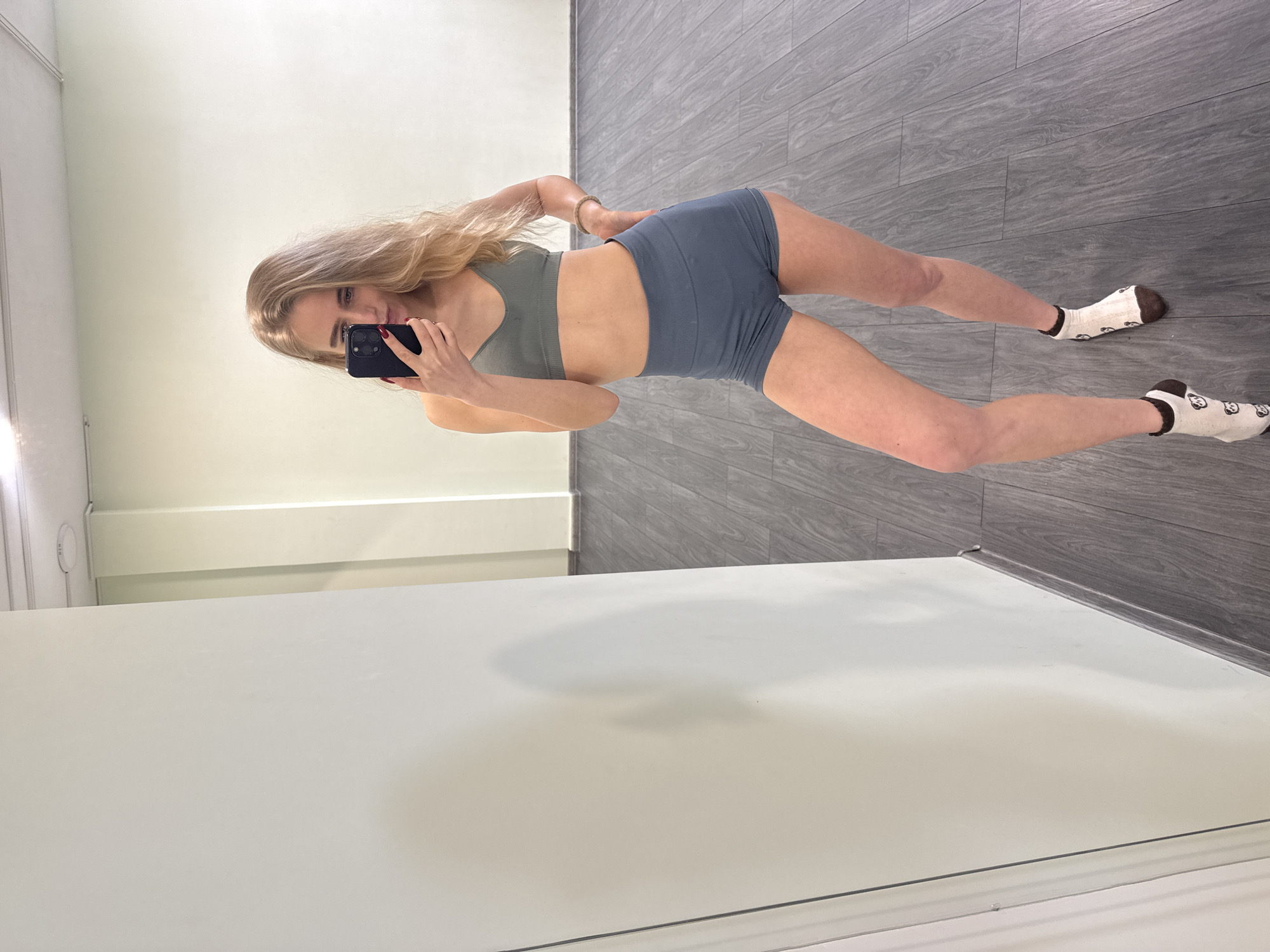 A young blonde woman in athletic wear is taking a selfie in a room with a white wall and dark gray wood-like flooring. She is wearing a gray sports bra and blue athletic shorts, along with socks patterned with cartoon faces. She holds a dark-colored smartphone in her left hand, her right hand resting on her hip. The room appears to be a workout space, potentially a gym or studio. The city of Unknown, Unknown is not evident from the visual information provided.