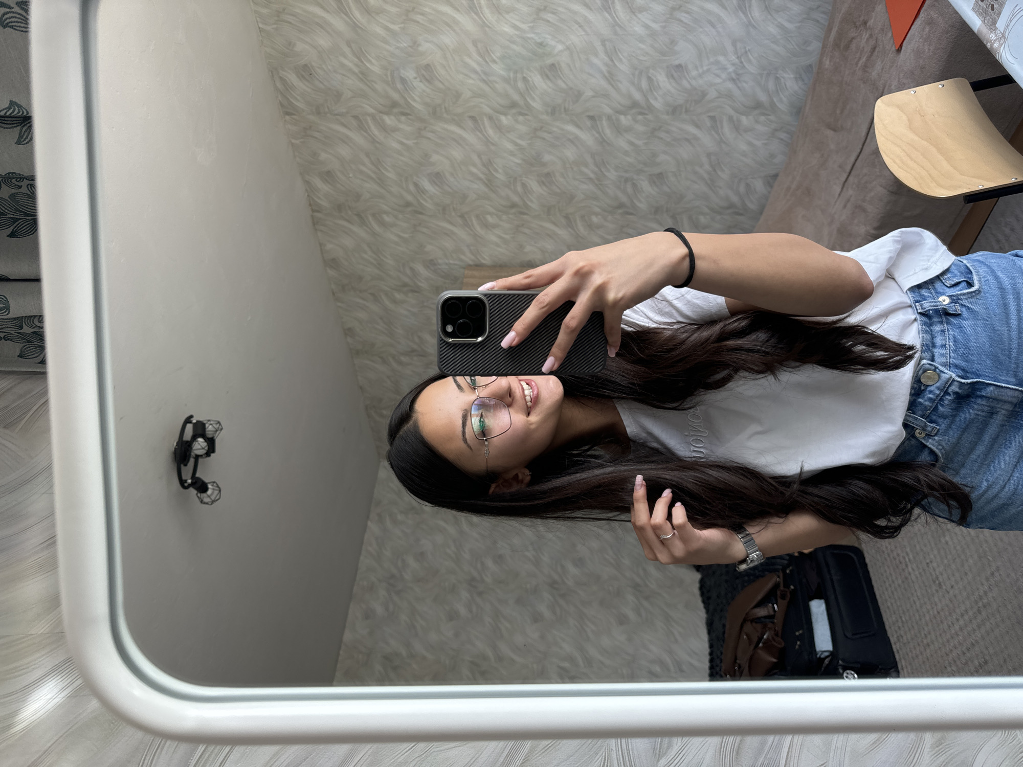 This media file captures a mirror selfie taken indoors in Sofia, Bulgaria. The central subject is a young woman, likely in her late teens or early twenties, with long dark hair, wearing glasses, a white t-shirt, and blue jeans. She is holding a smartphone, identifiable by its dark, textured case and triple camera system, to capture her reflection. She is smiling broadly, appearing happy and relaxed.
Notable details include a silver watch on her left wrist, a ring on her right ring finger, and a black elastic hair tie on her left wrist. The rectangular mirror has a thick white frame. The background reflected in the mirror features a wall with a distinct wavy, textured pattern, possibly wallpaper or carpet, in shades of grey and white. To the left of the woman, a light grey wall is visible with a black, industrial-style decorative light fixture mounted on it. To her right, parts of an upholstered furniture piece in a brownish-grey fabric are discernible, along with a wooden chair or stool with a curved seat, and a dark bag or suitcase on the floor. Faint white text, possibly "REVOLUTION" or a similar word, is visible on her white t-shirt. The lighting suggests it is daytime.
