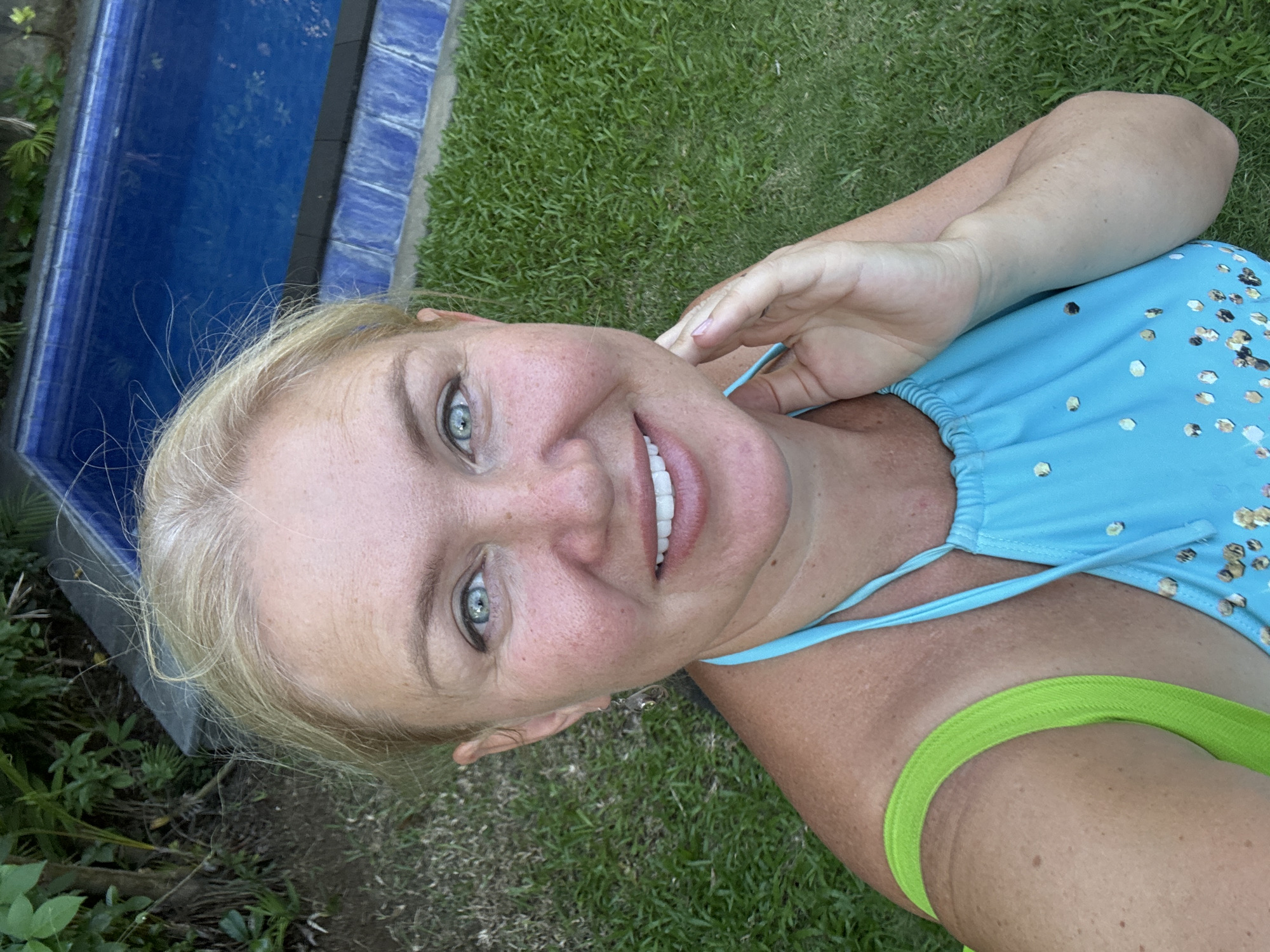 A woman with blonde hair and blue eyes lies on her side in the grass near a concrete structure with a blue surface. The woman is wearing a blue swimsuit with reflective gold circular adornments and a lime green strap is visible. The ground surrounding her is covered in green grass. The location appears to be an outdoor area within Kuta, Indonesia, indicated by the type of vegetation.