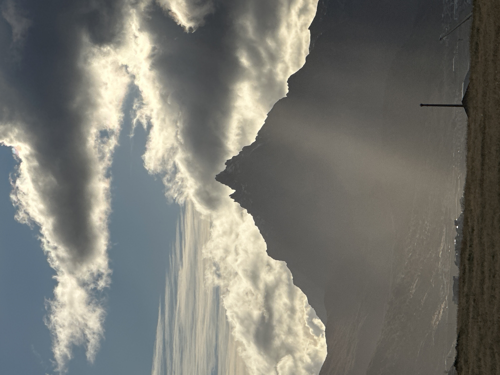 The image depicts a landscape with a prominent mountain range dominating the right side. Rain or sunlight is filtering down from the clouds onto the mountain. A telephone pole is visible in the right of the frame. To the left are streaky dark and light clouds with blue sky in the background. The scene suggests an open, potentially rural area on the outskirts of a settlement. It is not possible to verify that this photo was taken in Ushuaia.