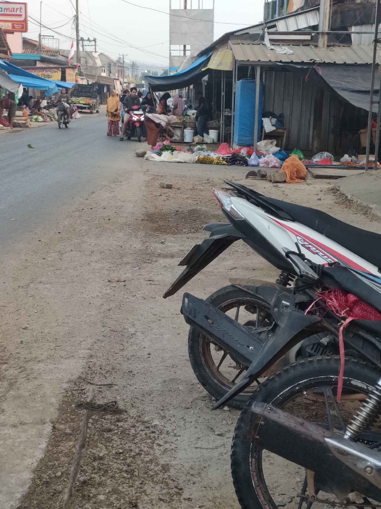The image depicts a street scene in Sukoyoso, Indonesia, likely a market area. In the foreground, two motorcycles are parked, their rear wheels and parts of their frames visible. The ground is a mixture of dirt and gravel, with a visible drainage channel. Beyond the motorcycles, a street is lined with market stalls under makeshift roofs. Several people are present, some walking and others on motorcycles. A white pickup truck is also visible. The stalls display various goods, including fruits like bananas and other produce, as well as sacks and bags. The architecture suggests a modest, possibly rural or semi-urban setting, with a mix of corrugated metal and tarpaulin structures. The overall impression is that of an active marketplace.