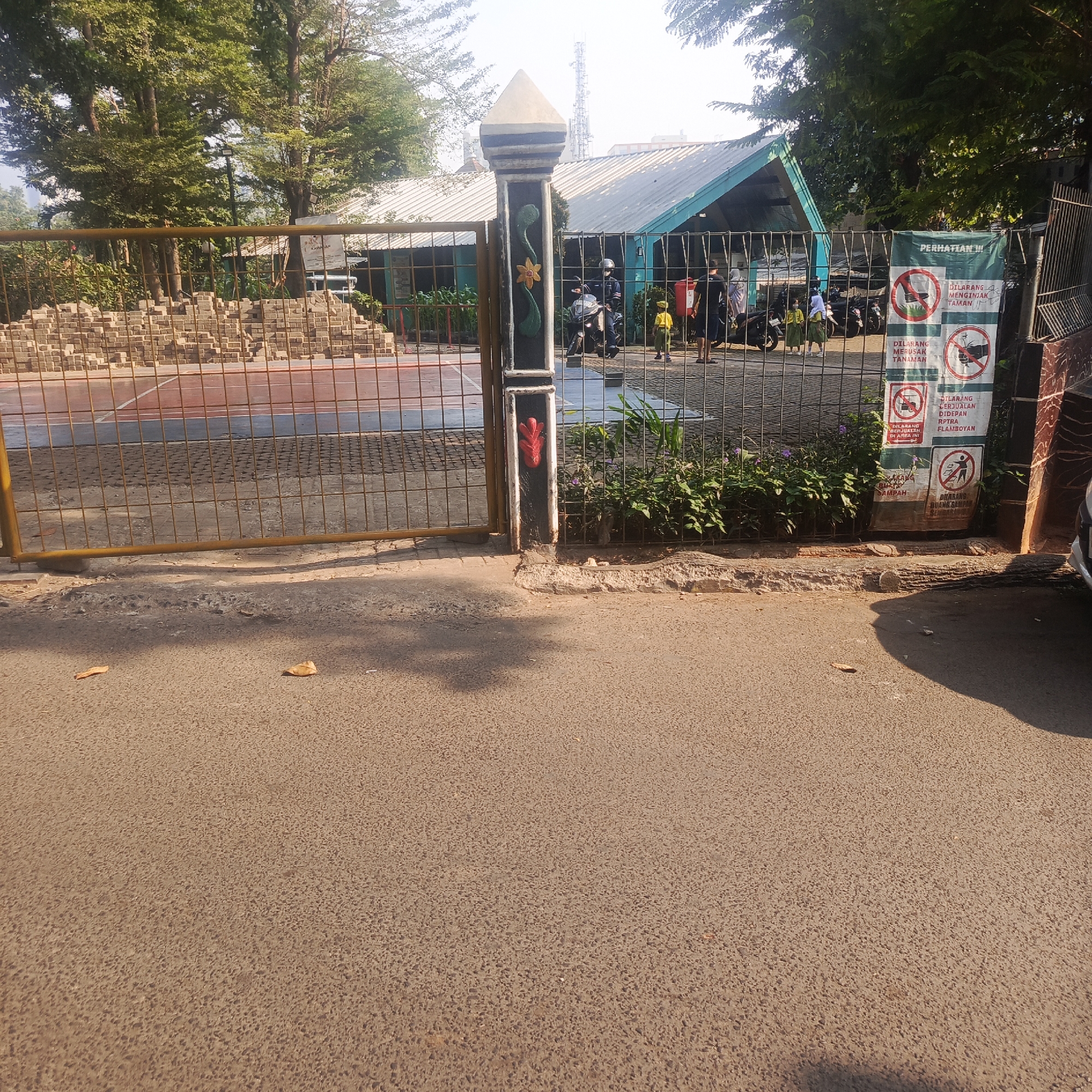 The image shows an outdoor scene with a yellow metal gate in the foreground, leading into what appears to be a courtyard or park area. Through the gate, a building with a light-colored roof and turquoise walls is visible. A pile of bricks is stacked to the left of the gate, and a paved court with red and white markings can be seen beyond. Several motorcycles are parked inside the gate, and a few people are present, including individuals wearing yellow uniforms, suggesting a school or organized group. A sign with multiple prohibition symbols and text in Indonesian is attached to the fence on the right side. The ground in the foreground is asphalt, with some scattered leaves. The environment appears to be a public or semi-public space, possibly a school campus or recreational facility, located in South Jakarta, Indonesia, given the language on the sign. The lighting suggests it is daytime, with sunlight casting shadows on the ground.