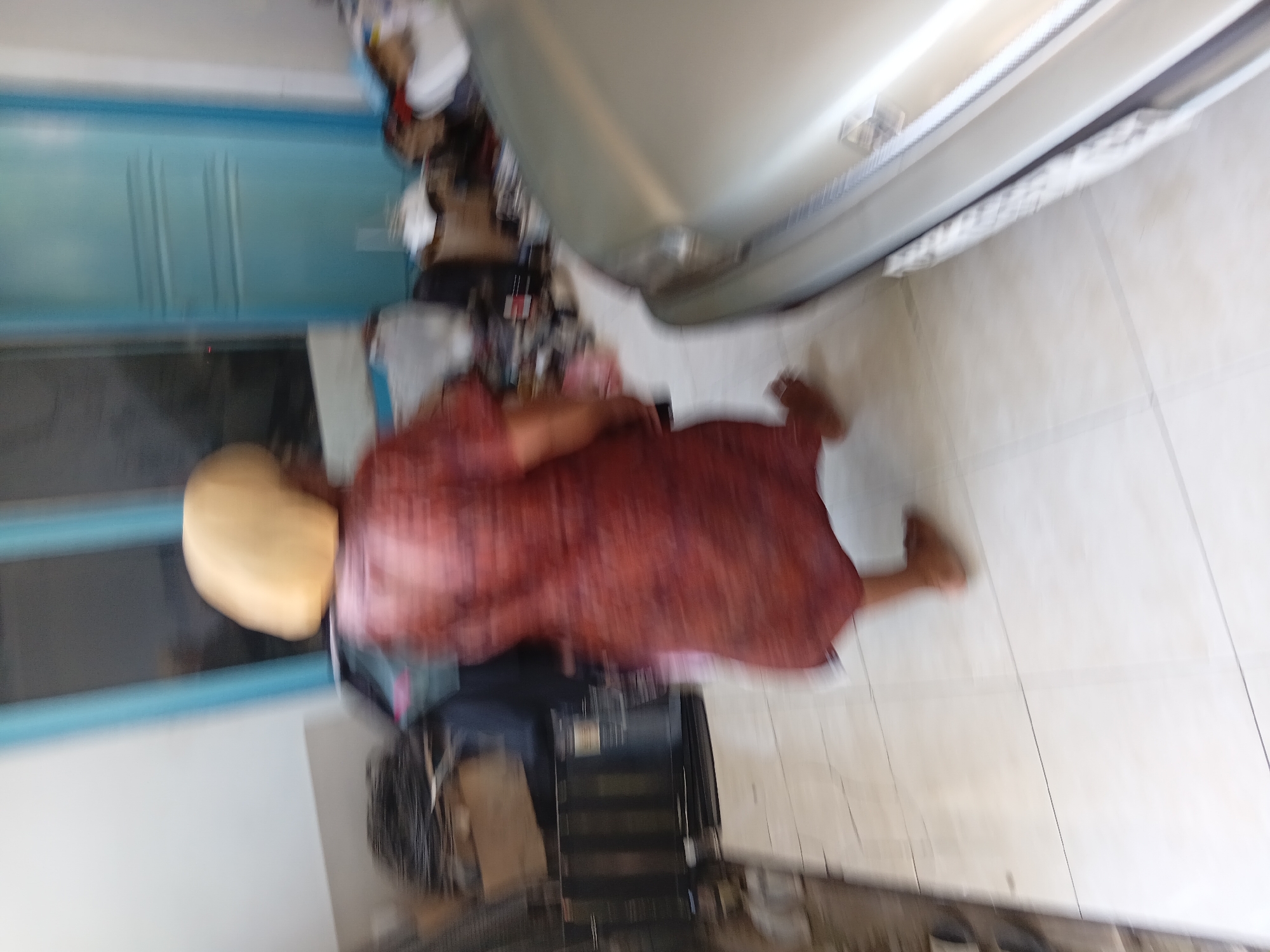 A person with a blonde bob haircut, wearing a red patterned dress, is walking across a tiled floor. The person is facing away from the camera. To the right of the person is the front of a silver car. In the background, there are blue doors and various objects that appear to be stored or discarded, including what looks like a black fan. The scene is blurry, suggesting motion. This image was taken in Tanggul Wetan, Indonesia.