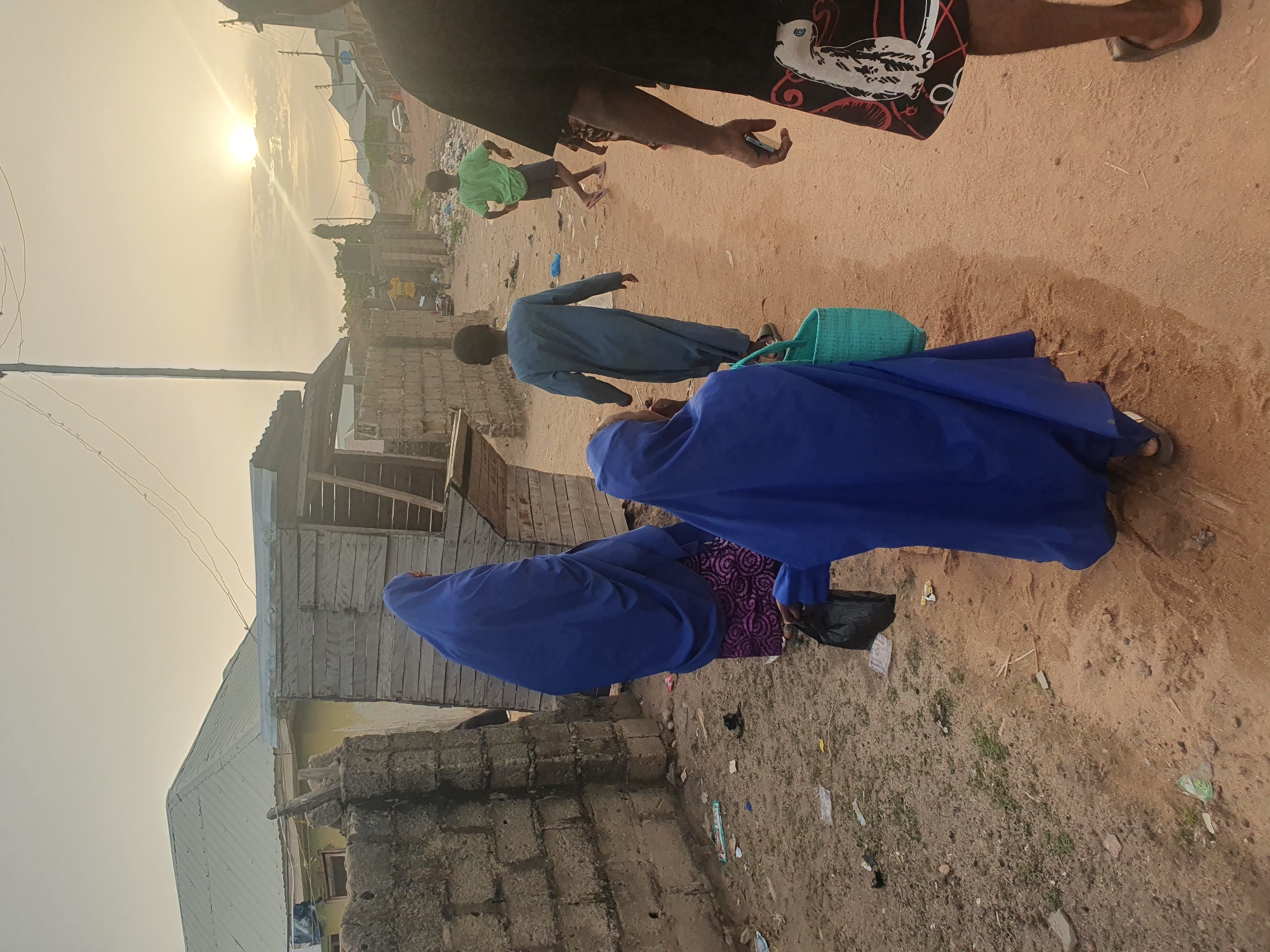 The image shows a street scene in Lokoja, Nigeria. The setting appears to be a low-income residential area. In the foreground, two women are walking away from the camera. Both are wearing blue garments. The first woman also has a black bag and a patterned purple fabric underneath her blue cloth. The second woman is walking closely behind the first and has a turquoise colored bag. Further ahead, a man in a light blue robe walks. A child in a green shirt and black shorts runs in the distance to the left. A man stands in the very foreground of the image. The terrain is sandy. There are buildings made of blocks with metal roofs to the left. There is trash scattered on the ground. The sky is bright with the sun setting in the background.