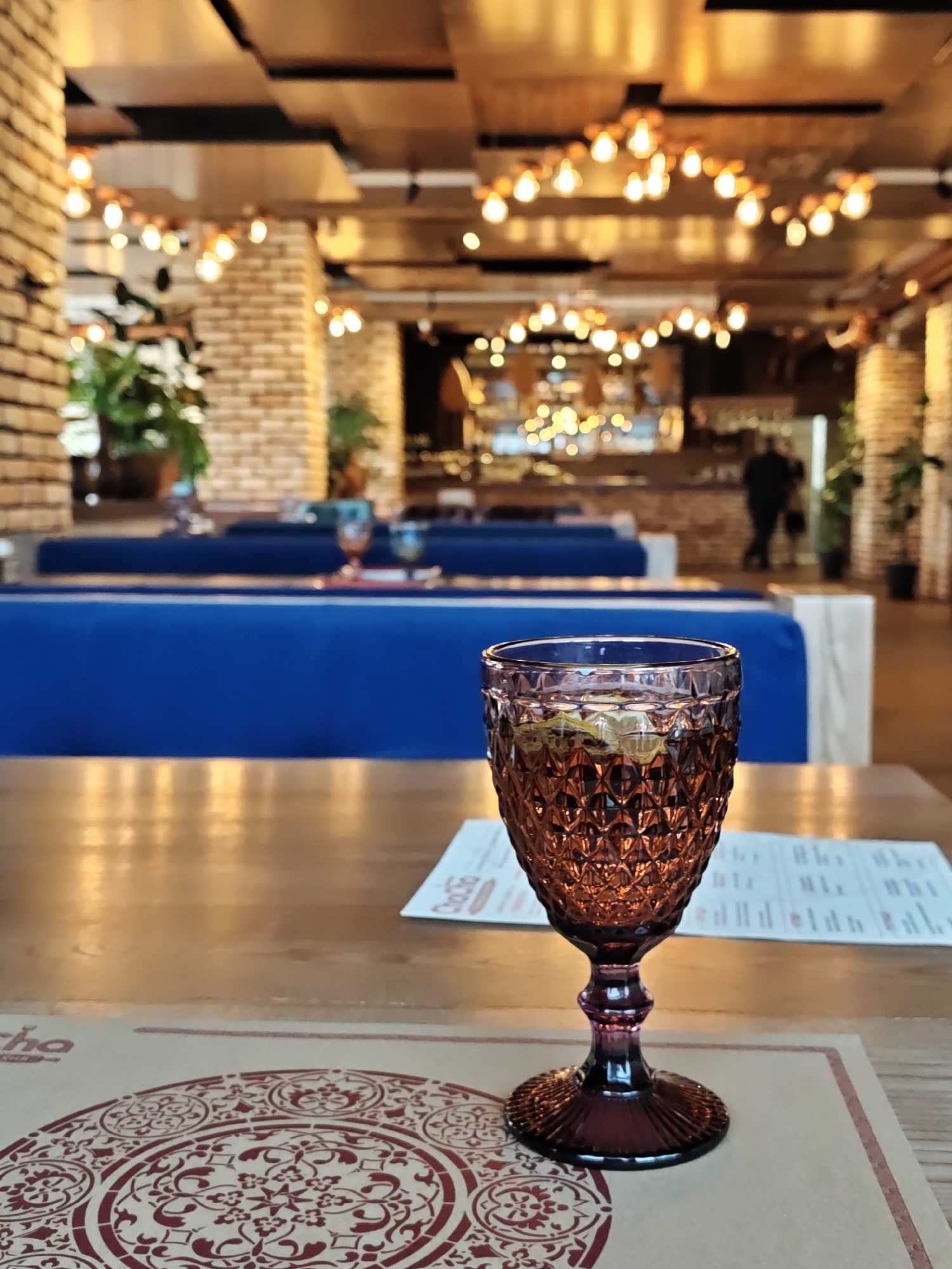 The photo shows a wine glass on a table in a restaurant in Vinnytsia, Ukraine. The glass is purple and faceted, filled with a light-colored liquid, and sits on a decorative placemat with a red and white circular pattern. Behind the glass is a menu. In the background, there are blue upholstered booths, exposed brick columns, and a bar area with people. The ceiling has wooden panels and decorative light fixtures.