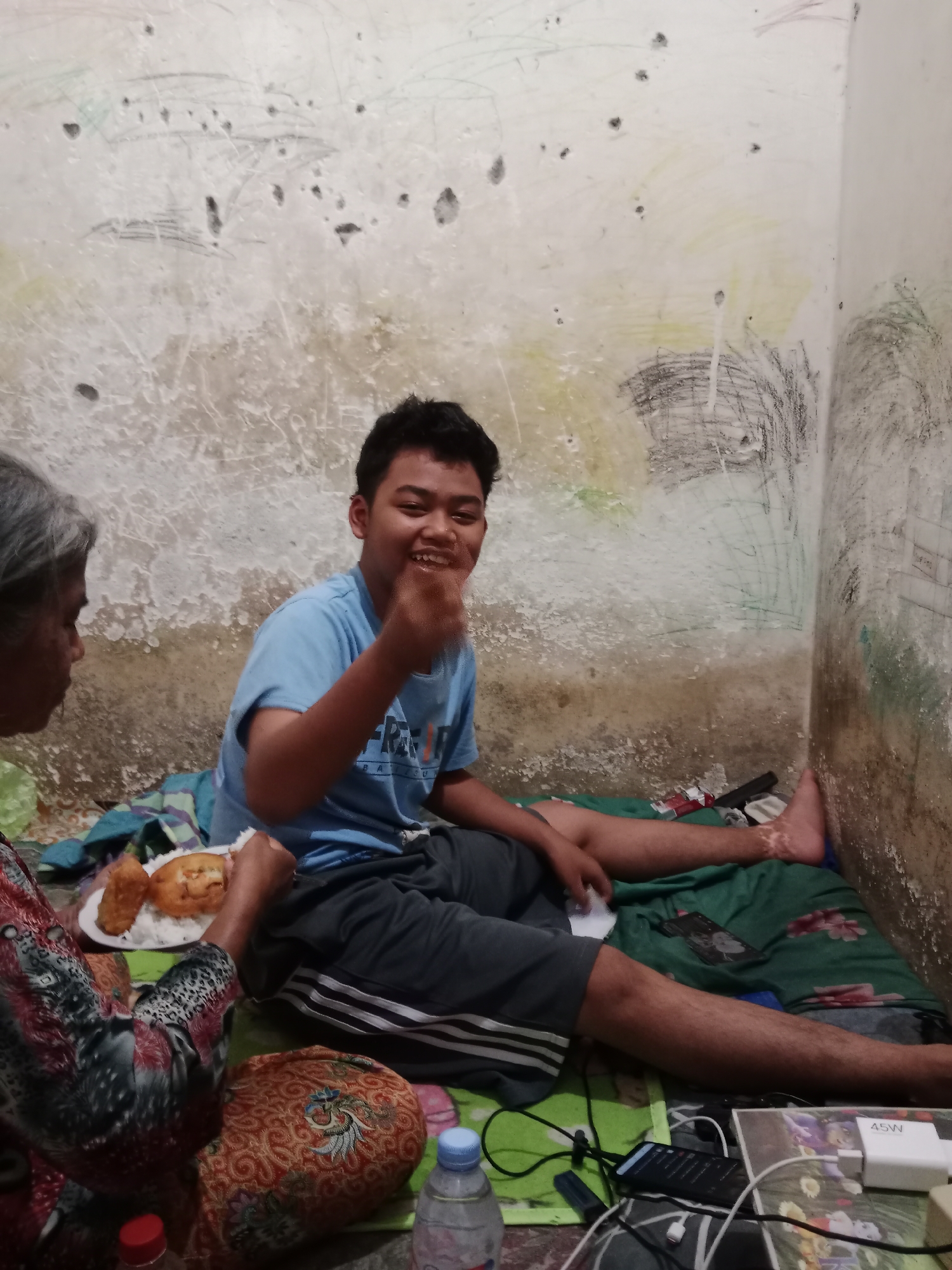In a dimly lit room with heavily textured and marked walls, two individuals are seated on the floor, which is covered with a green blanket and patterned fabrics. To the left, an elderly person with gray hair, wearing a patterned top, is holding a plate of rice and fried food. To the right, a young male with short dark hair, wearing a light blue t-shirt and dark shorts, is smiling and gesturing with his right hand, which appears to have food on it. He is seated on the green blanket, with his legs extended. Various objects are scattered around them on the floor, including cigarettes, a mobile phone, charging cables, a white power adapter, and plastic bottles, one of which is a clear bottle of water. The overall impression is a casual meal shared in a modest indoor setting. The location is identified as Ringinrejo, Indonesia.