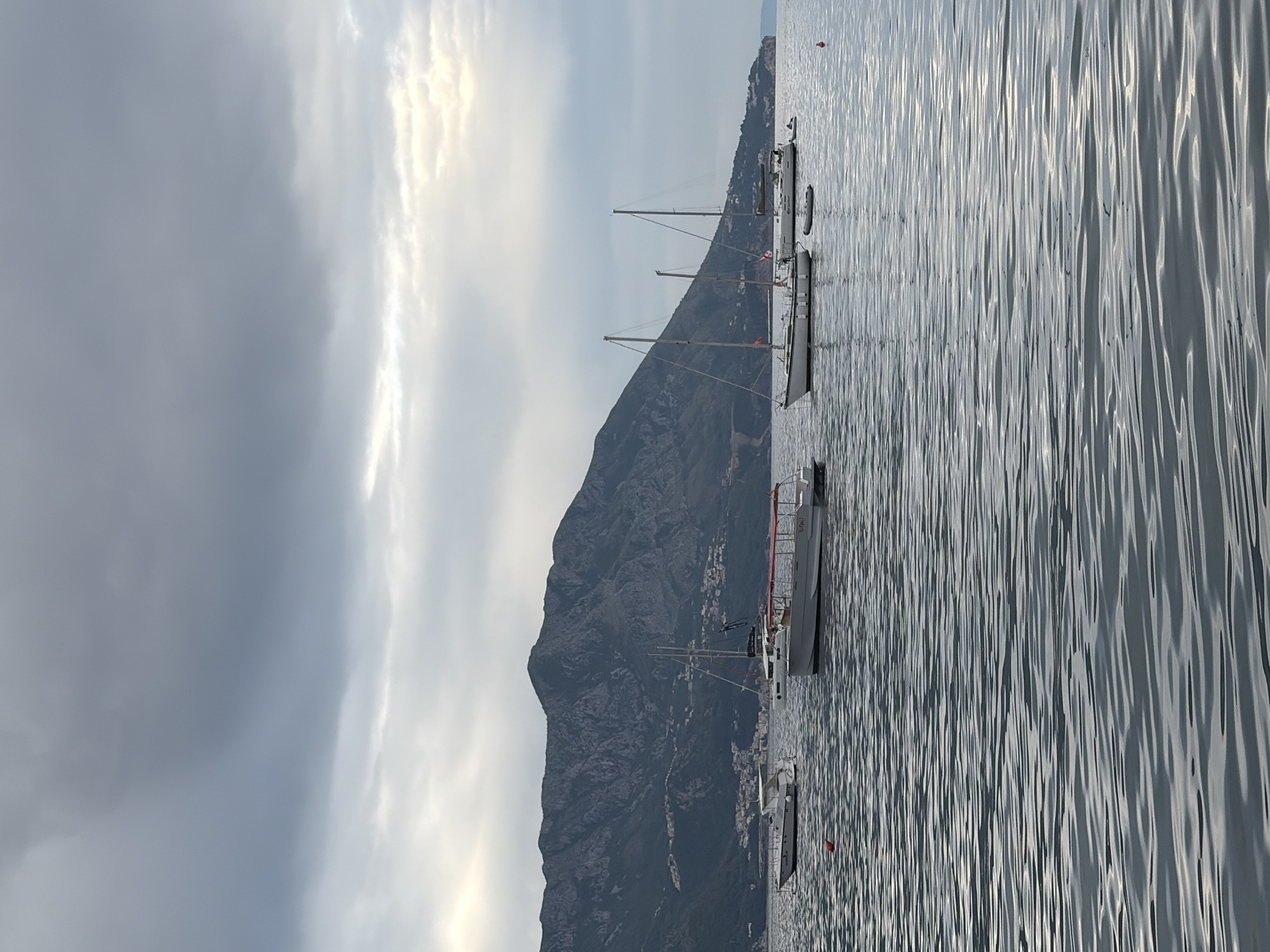 The image depicts a waterfront scene in Budva, Montenegro. Several sailboats are docked or moored in the calm water near the shoreline. A mountainous landscape rises behind the buildings and coastal areas along the water's edge. The sky is overcast with dense cloud cover. Ripples are present on the water's surface, reflecting light.