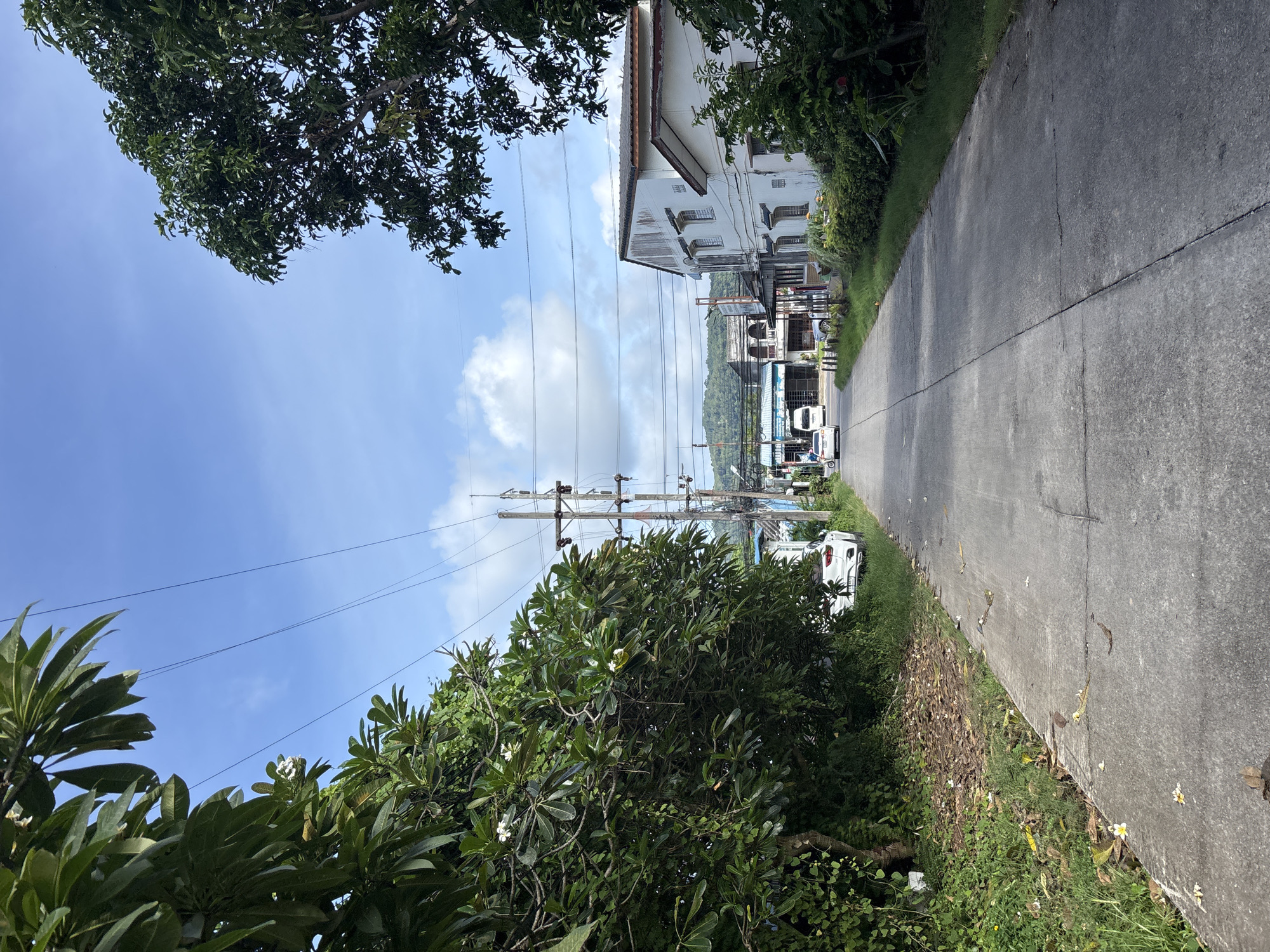 The image depicts a paved road in Chalong, Thailand, lined with lush green trees on the left and buildings on the right. The sky is a vibrant blue with scattered white clouds. Power lines are visible overhead, supported by utility poles. Several vehicles are present, including a white SUV parked on the side of the road and two vans driving away from the camera. The buildings appear to be a mix of residential and commercial properties, with some showing signs of age and wear. The overall scene suggests a somewhat rural or suburban area within Chalong, characterized by natural vegetation and everyday infrastructure.
